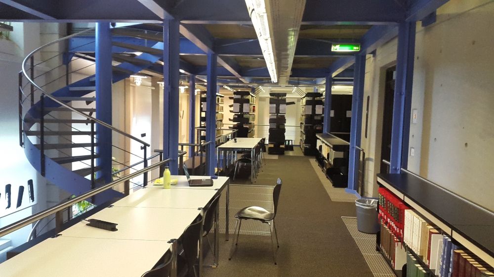 ZUB-1, Bibliothek: Sicht von der linken Seite des Stockwerkes aus auf den nur mit Schlüssel zugänglichen Eingang auf der rechten Seite, auf die 8 Arbeitsplätze und die Wendeltreppe links, die hinauf zum 2. Obergeschoss oder hinab zum Erdgeschoss führt.