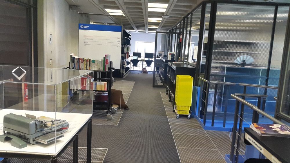 ZUB-2, Bibliothek: Sicht auf den Eingang links und die Wendeltreppe rechts, die zu den anderen Bibliotheksstockwerken hinunter führt. Geraudeaus hinten befindet sich die Selbstausleihstation links und eine der Recherchestationen rechts. Daneben befindet sich der Rückgabewagen.