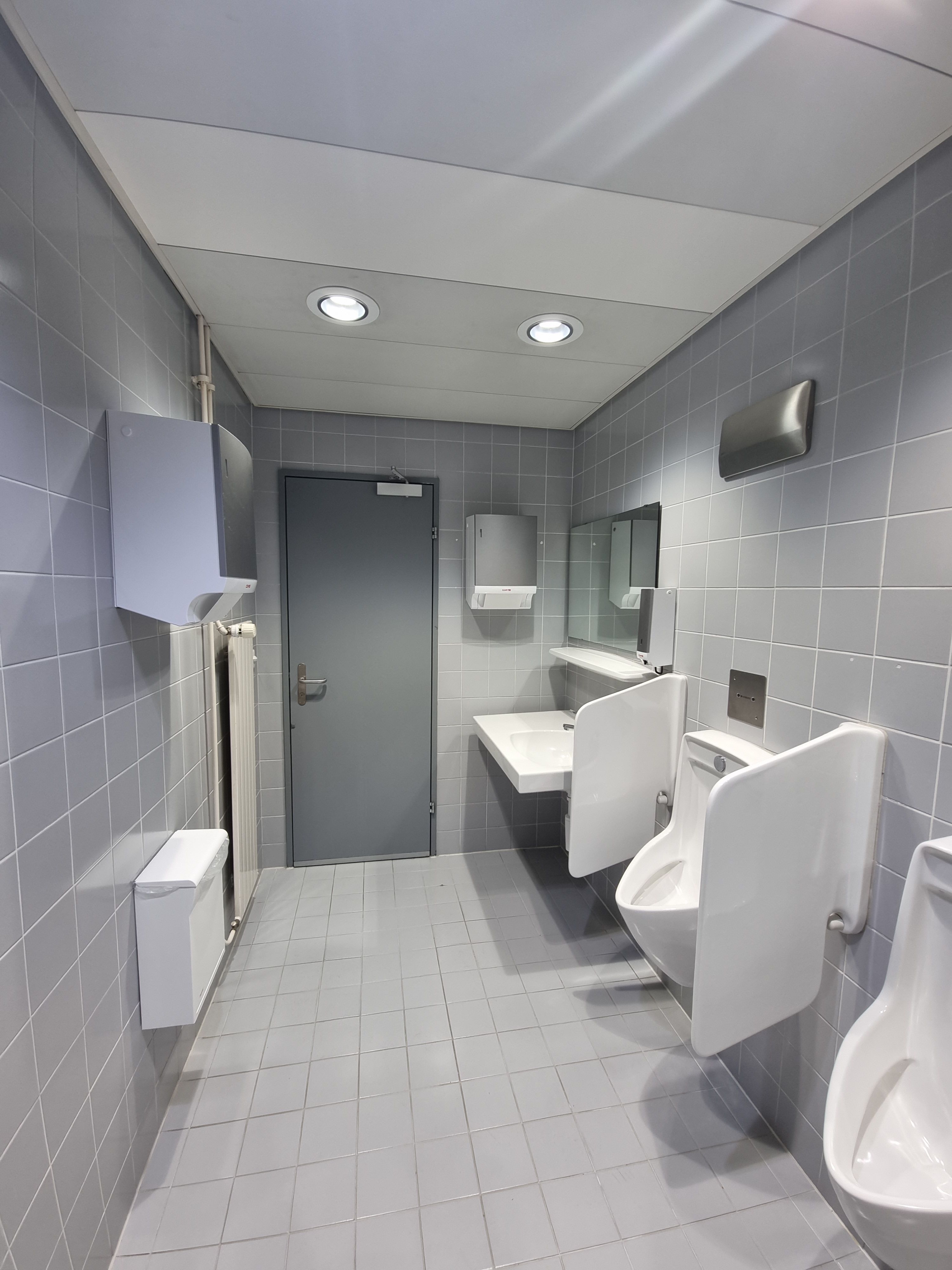 Da 714: Bild des WCs von der Wand gegenüberliegend des Eingangs aus. Auf der rechten Seite sind zwei Pissoirs und ein Waschbecken. Auf der linken Seite an der Wand angemacht ist ein Stoffhandtuchspender. Links vom Waschbecken aus an der Wand befindet sich ein weiterer Stoffhandtuchspender.