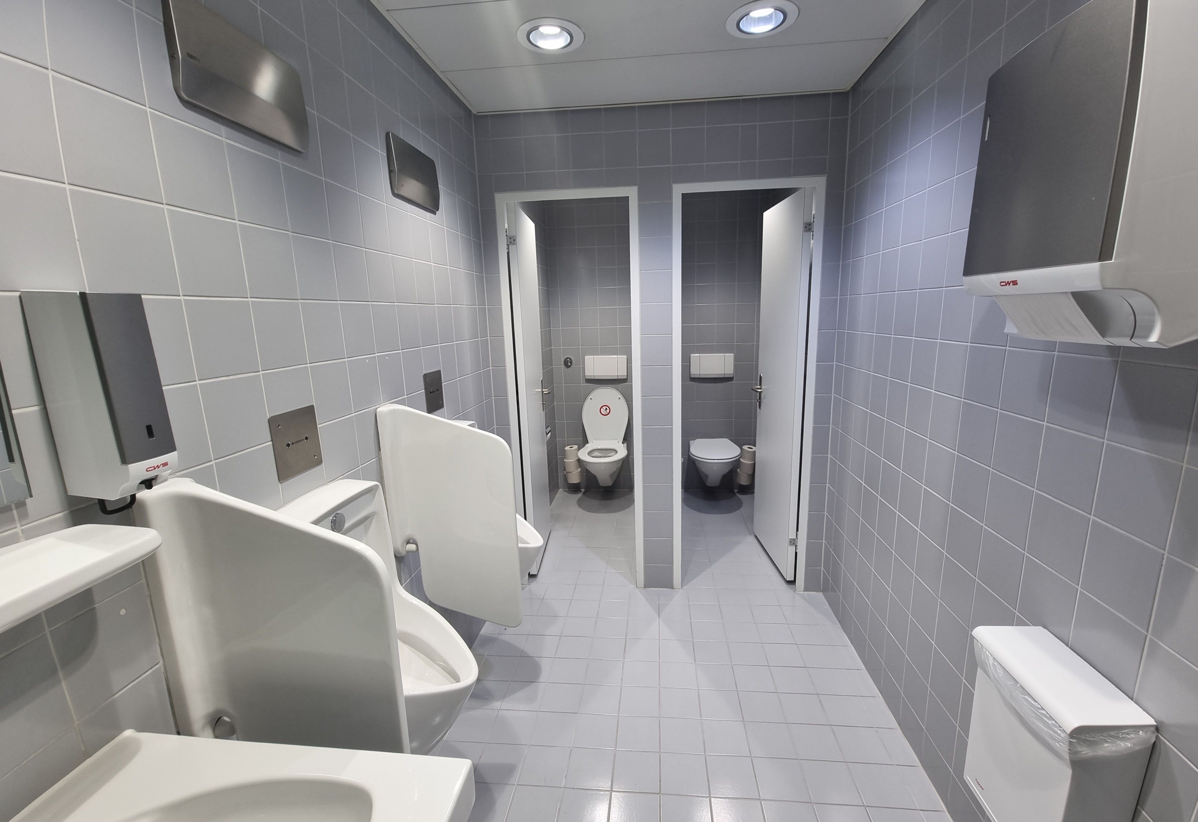 Da 714: Bild vom WC mit dem Blick vom Eingang aus. Auf der linken Seite befinden sich das Waschbecken und zwei Pissoirs. Auf der rechten Seite ist der Stoffhandtuchspender und ein geschlossener Abfallkorb. Geradeaus sind zwei Toilettenkabinen.