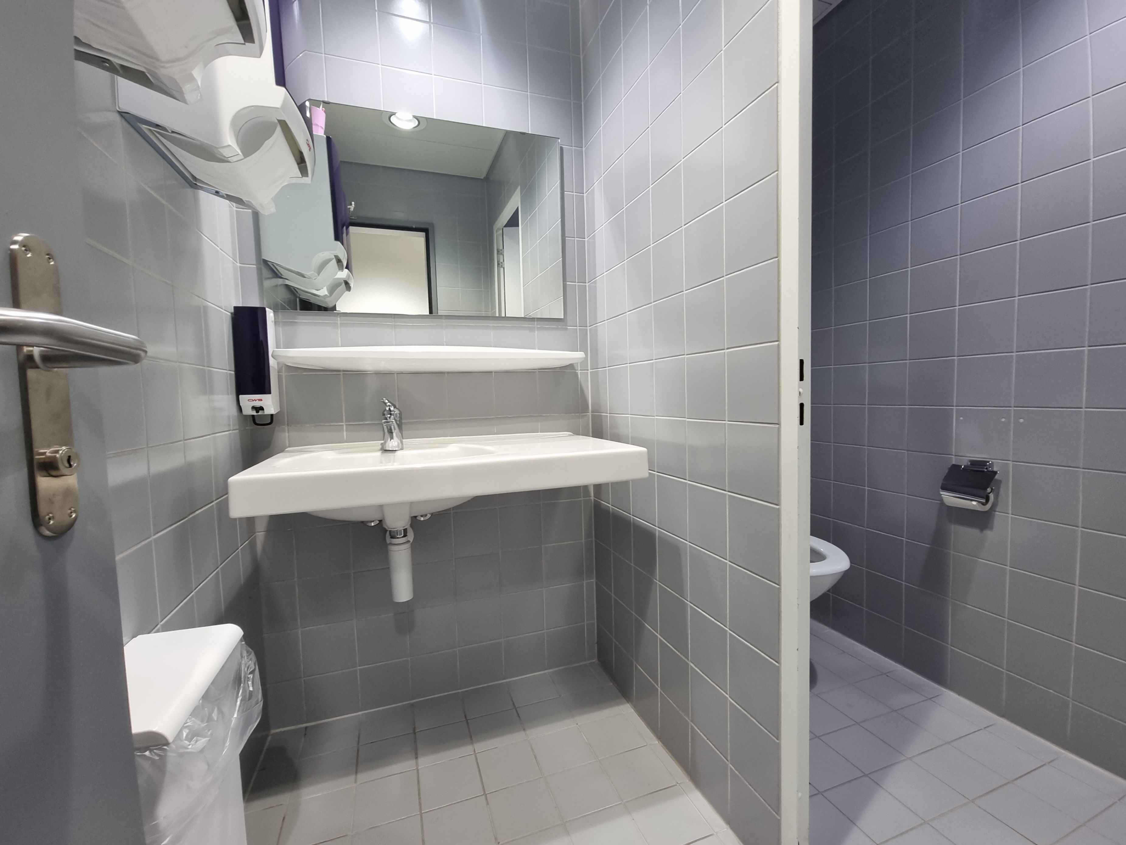 Da 726: Ein Bild des WCs vom Eingang aus. Das Waschbecken ist geradeaus vom Eingang. Links vom Waschbecken an der Wand hat es zwei Stoffhandtuchspender und einen geschlossenen Abfall. Auf der rechten Seite befindet sich die Toilettenkabine.