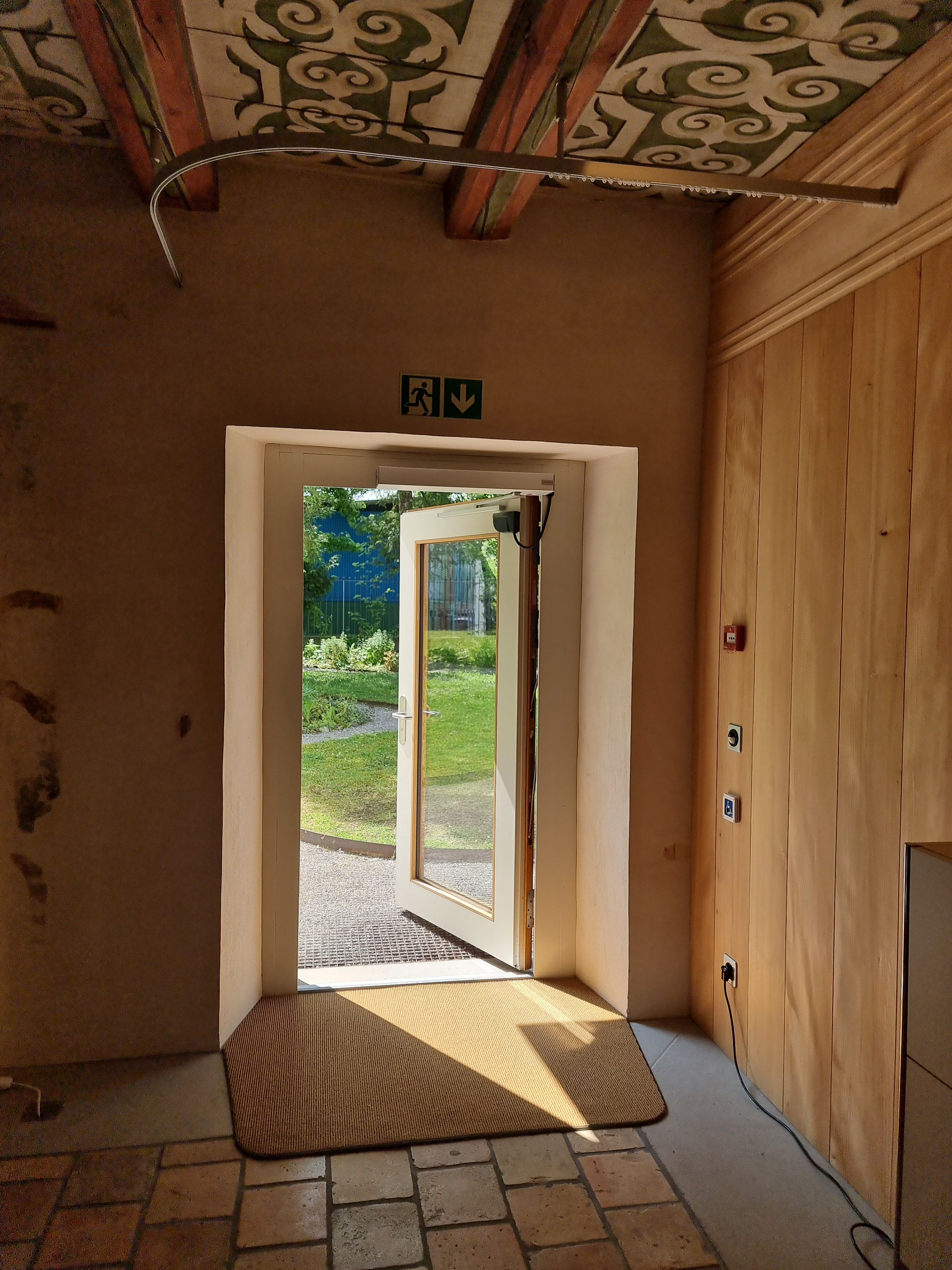 SOB Gebäude Erdgeschoss: Auf dem Bild ist die barrierefreie Tür von Innen abgebildet während des Zufallens. Oberhalb der Tür ist das Zeichen zur Markierung eines Notausgangs abgebildet. Die Tür ist automatisch und grösstenteils durchsichtig verglast.