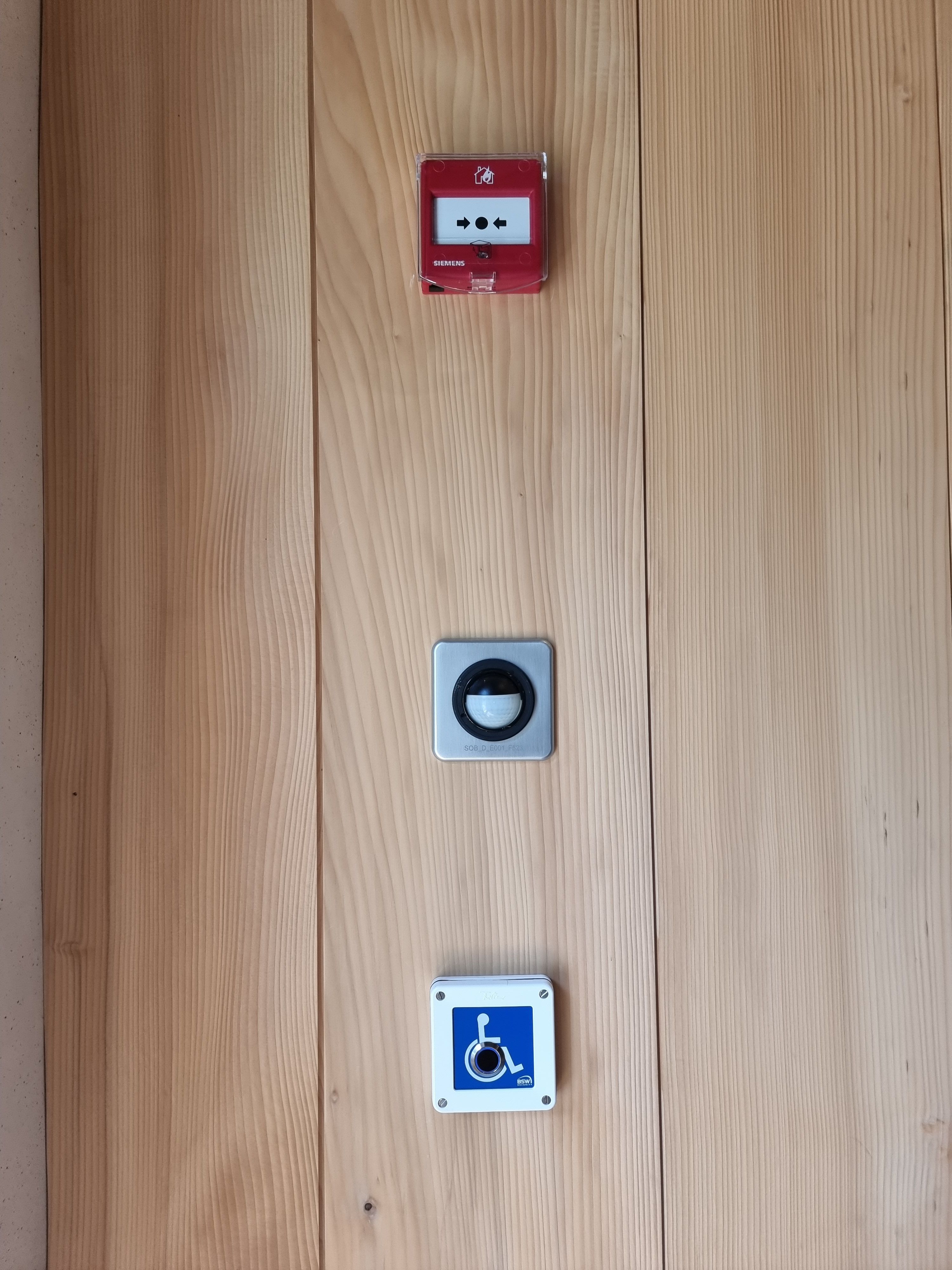 SOB Gebäude Erdgeschoss: Auf dem Bild sind die Elemente rechts neben dem barrierefreien Ausgang abgebildet. Darauf sieht man eine der Brandmeldeanlagen des Stockwerks. Der Brandmelder muss nicht eingeschlagen werden. Darunter ist ein Knopf markiert mit einem Rollstuhlzeichen. Der Knopf betätigt die automatische Türe für den barrierefreien Eingang. Der Knopf muss nicht gedrückt werden. Es ist ein Sensor.