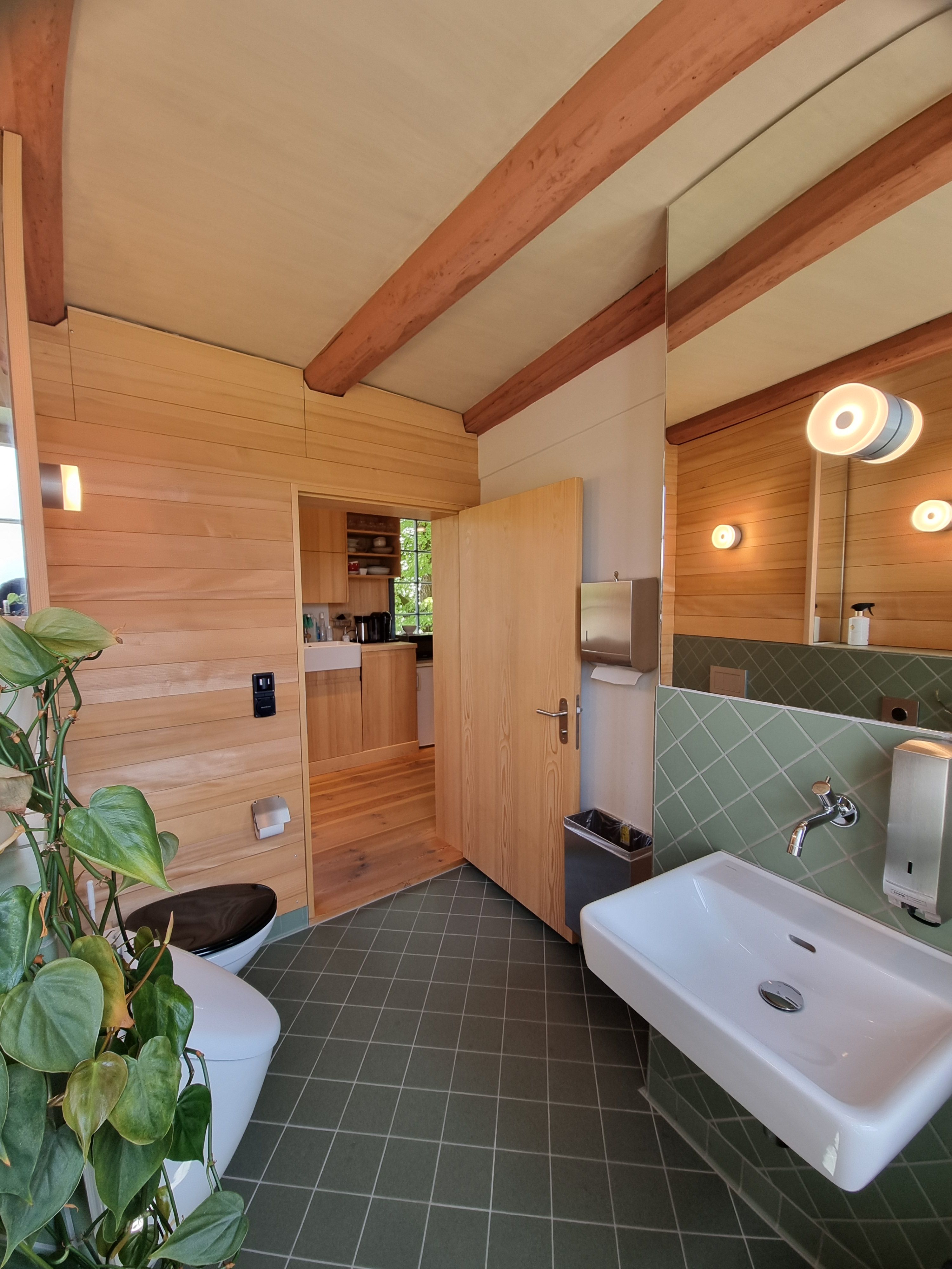 F-108: Bild des WCs mit dem Blick vom Fenster auf die WC-Tür. Man sieht die 4cm hohe Türschwelle. Etwas links ums Eck neben dem Waschbecken befindet sich der Handtuchspender. Unter dem Handtuchspender hat es einen offenen Abfallkorb. Der Abfallkorb ist an der Wand montiert.