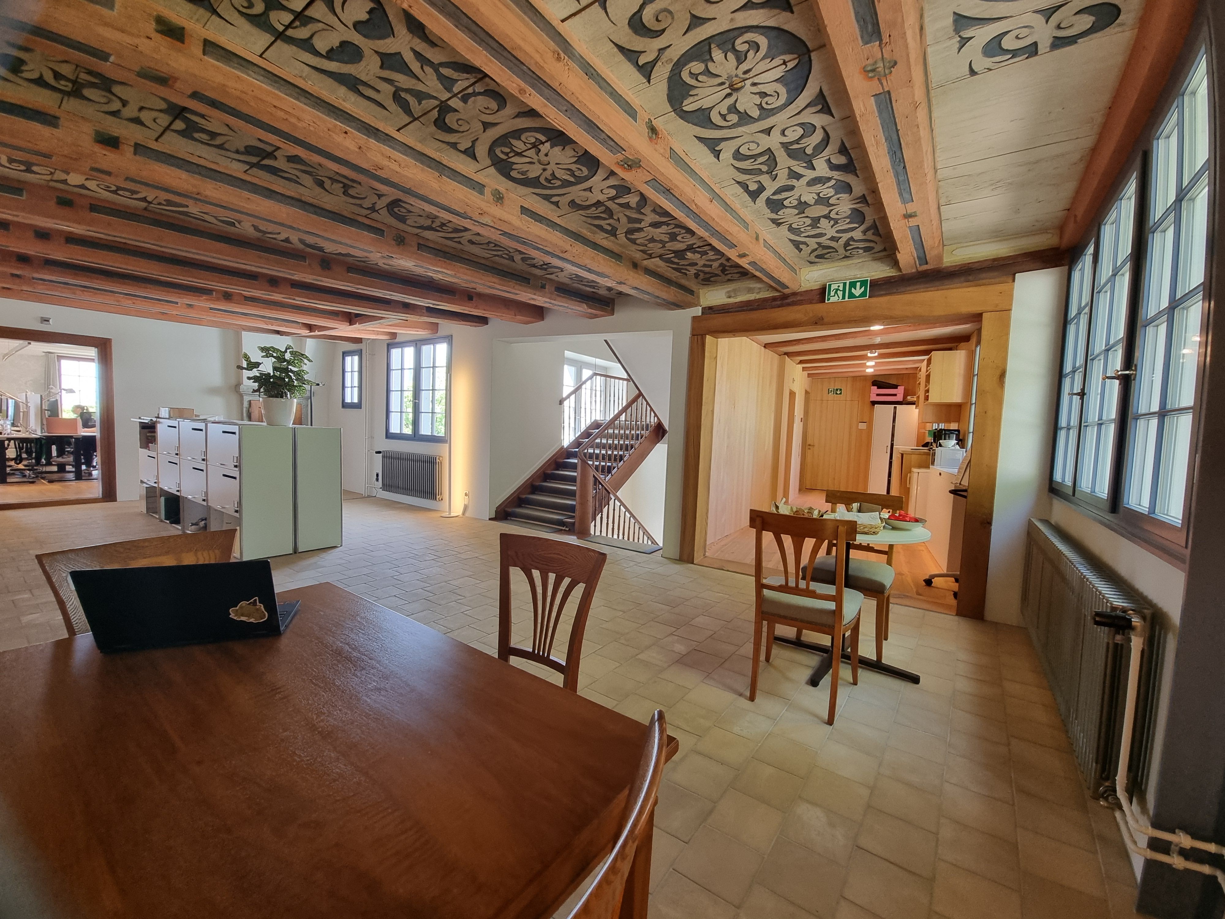 SOB 1. Stock: Bild des Stockwerks mit Sicht auf den Fluchtweg und die Treppe. In der Mitte des Raums vor dem Fluchtweg befinden sich ein Tisch und zwei Stühle.