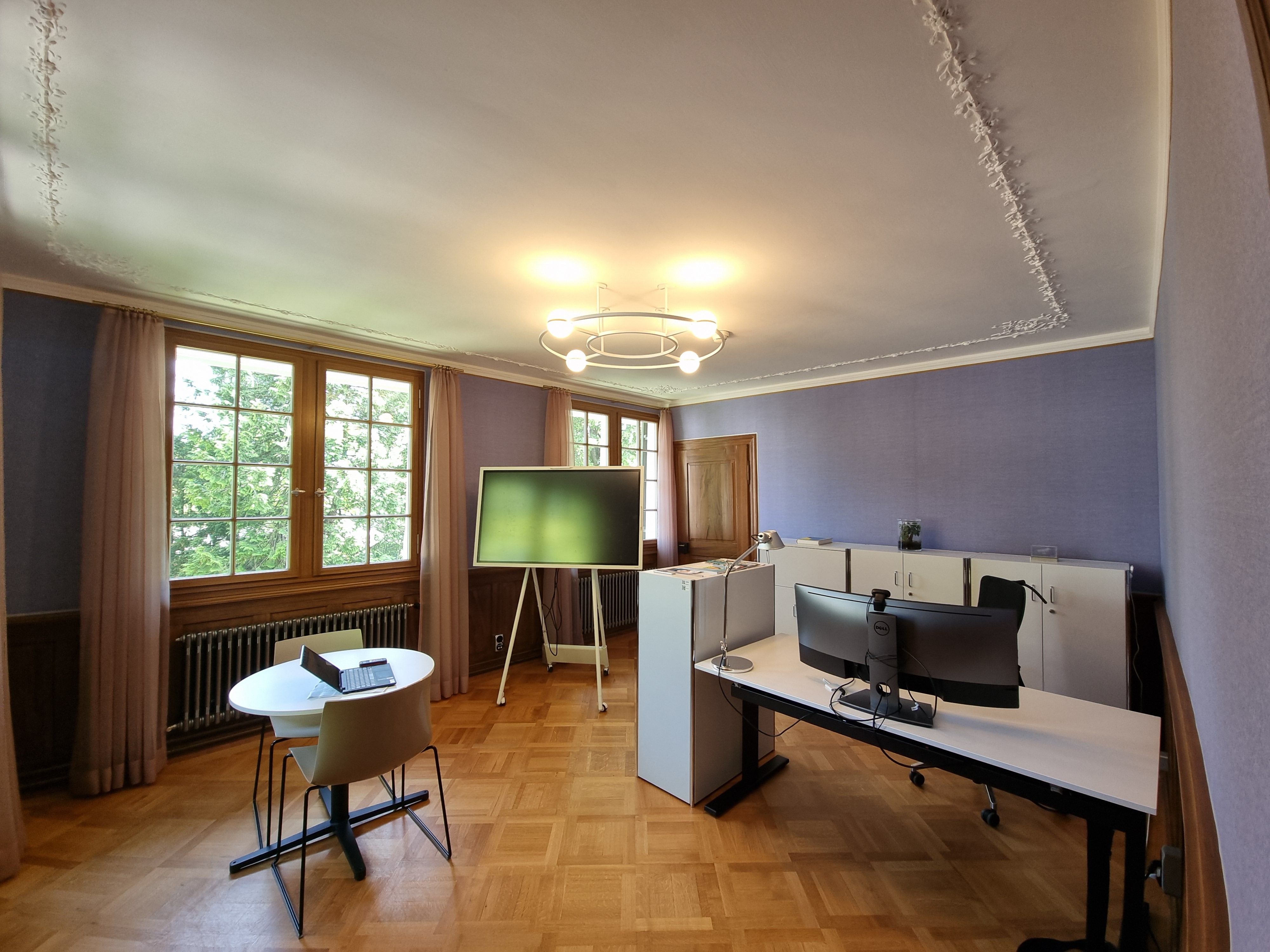 G-102: Bild des Besprechungszimmers von der Tür aus. Die Wand hat eine lila Tapete. Im Raum befindet sich ein grosser Bürotisch und ein kleiner runder Tisch mit zwei Stühlen. Zwischen dem kleinen Tisch und dem grossen Bürotisch steht ein grosser Monitor.