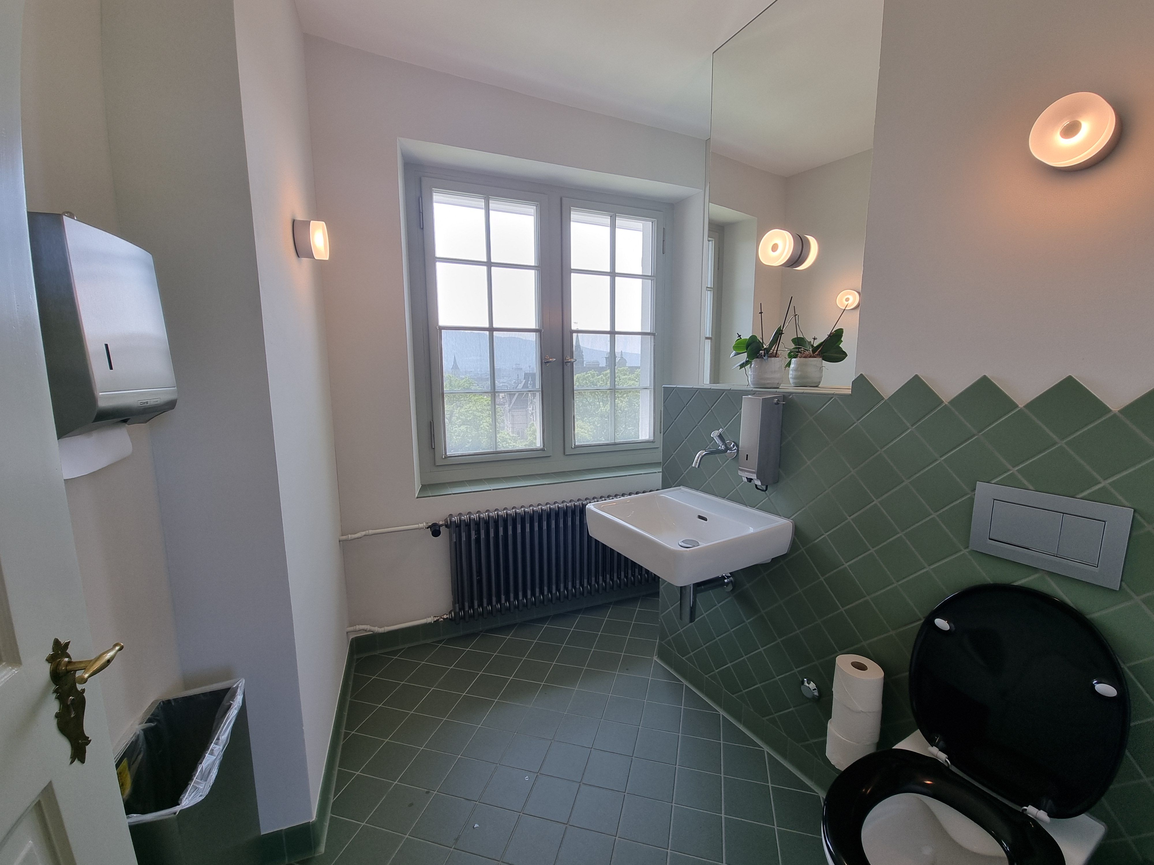 G-108: Bild des WCs von der Tür aus betrachtet. Das Licht ist an. Auf der rechten Seite befindet sich das Sitzklo. Links davon ist das Waschbecken. Rechts vom Waschbecken befindet sich ein grosses Fenster, das man aufmachen kann. Gegenüber des Sitzklos befindet sich der Handtuchspender und ein offener Abfallkorb darunter.