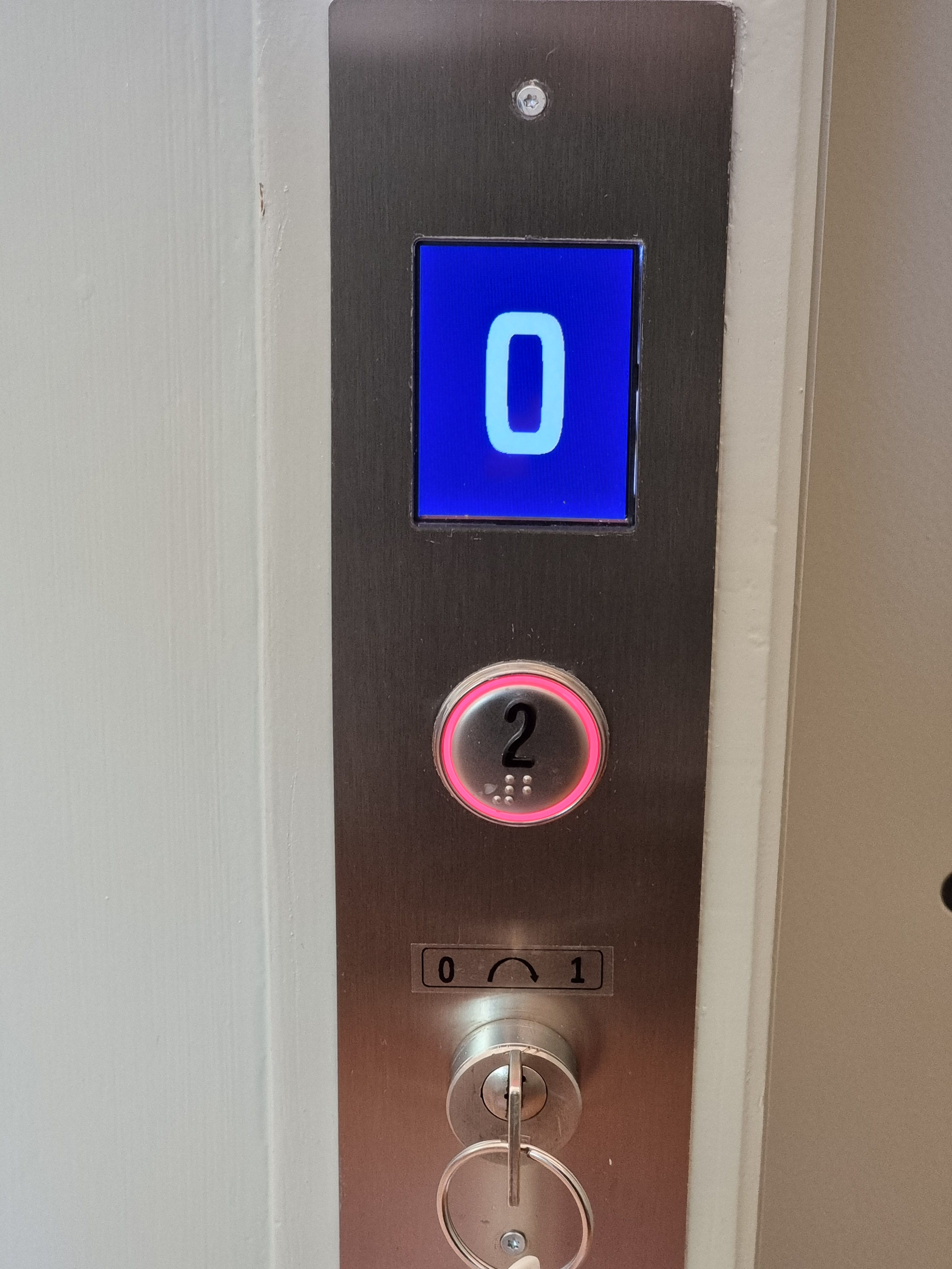 E-400J: Bild vom Bedienungsfeld ausserhalb vom Lift. Der Eurokey ist unter der Lifttaste eingesteckt. Die Taste ist beschriftet mit der Nummer 2. Der Rand der Lifttaste leuchtet rot. Oberhalb der Taste befindet sich ein rechteckiges Display. Auf dem blauen Hintergrund des Displays sieht man die Zahl 0 in weisser Schriftfarbe. Der Lift befindet sich auf dem Stockwerk 0.