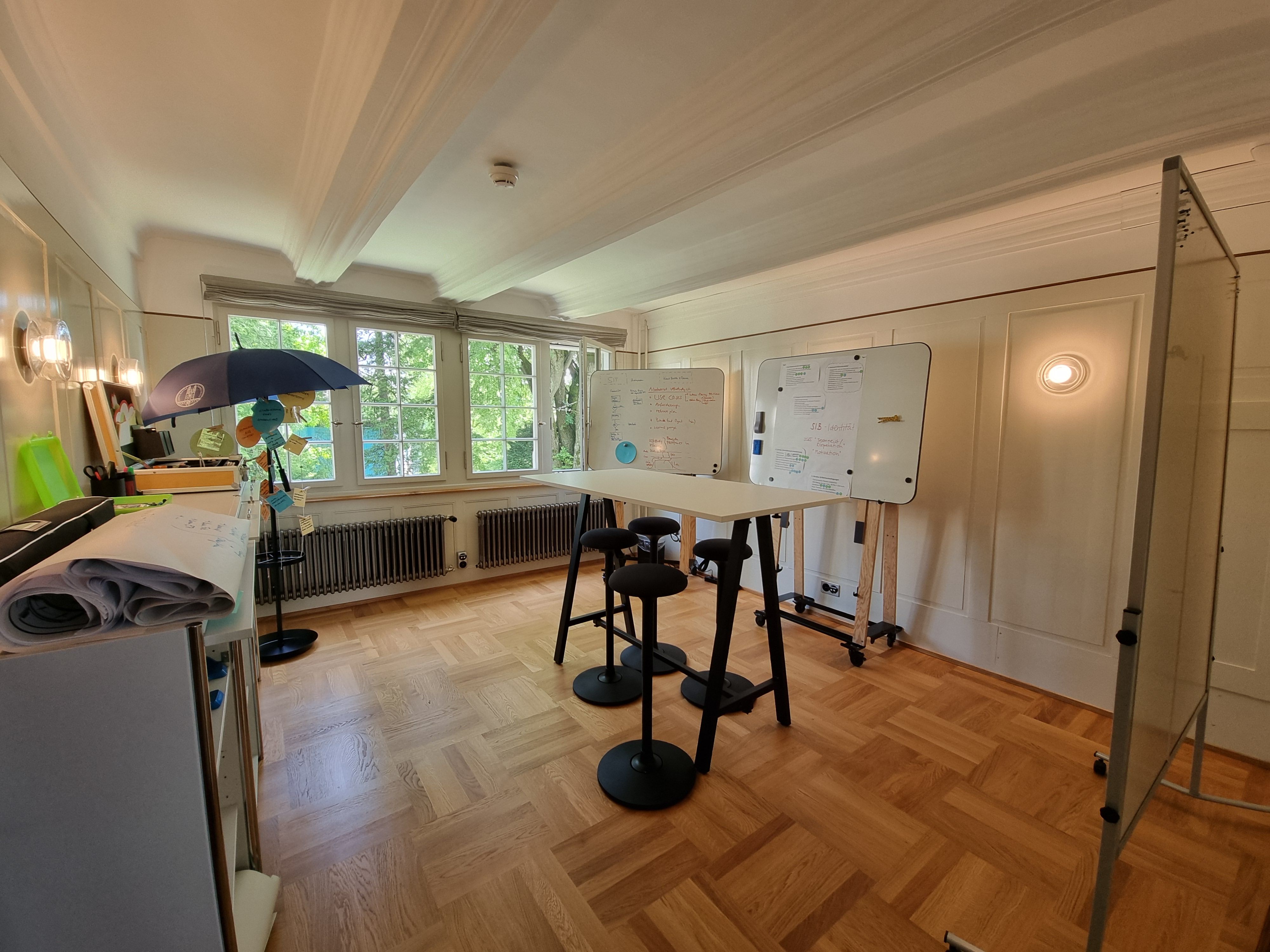 F-103: Blick auf den Workshopraum von der Tür aus. In der Mitte des Raums hat es einen hohen Tisch und hohe Stühle rundherum. Im Raum verteilt befinden sich drei grosse Whiteboards. Auf der linken Seite des Zimmers befindet sich eine längliche Kommode mit Büromaterial. Alle Fenster befinden sich an der Wand gegenüber des Eingangs.