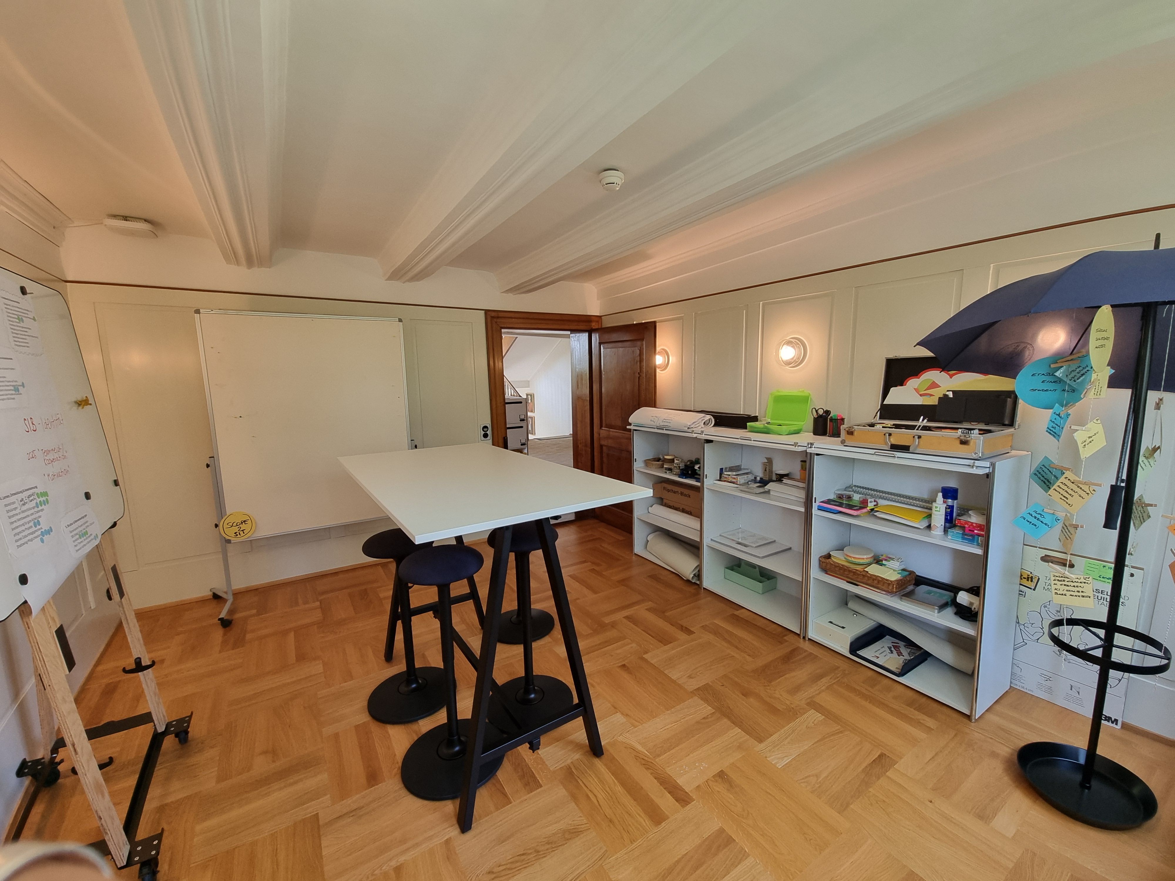 F-103: Bild des Workshopraums vom Fenster aus. Die Eingangstür ist offen. Auf der rechten Seite sieht man die längliche Kommode mit Büromaterial. Auf der linken Seite sieht man die Whiteboards. In der Mitte befindet sich ein hoher Tisch mit hohen Stühlen rundherum.