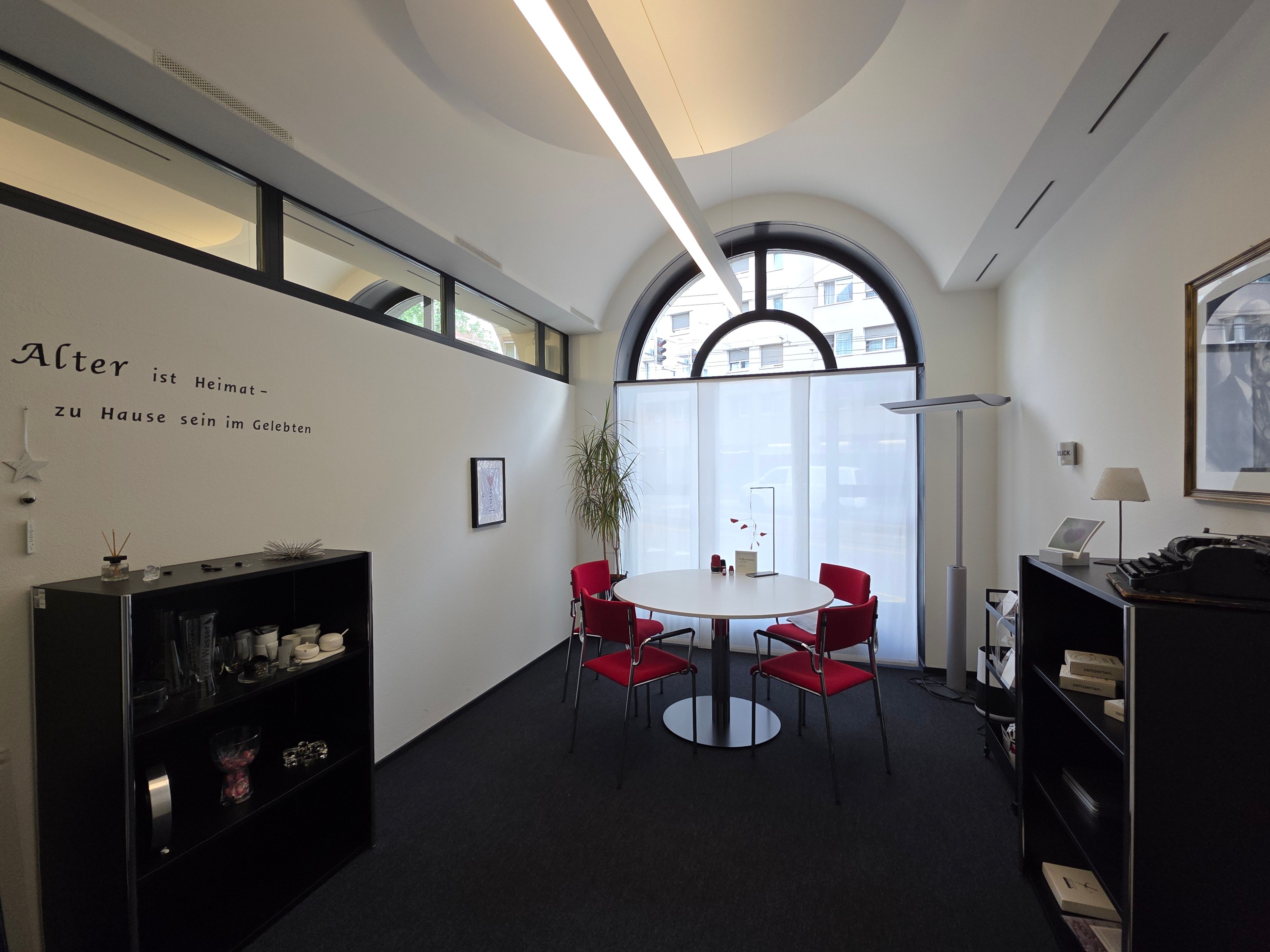 Beratungszimmer E-005: Bild vom Beratungsraum von der Eingangstür aus. Im Zimmer hat es einen runden Tisch mit vier roten Stühlen rundherum. Links und rechts im Raum befinden sich zwei schwarze Regale. Die Wand gegenüber der Tür hat ein grosses Fenster.