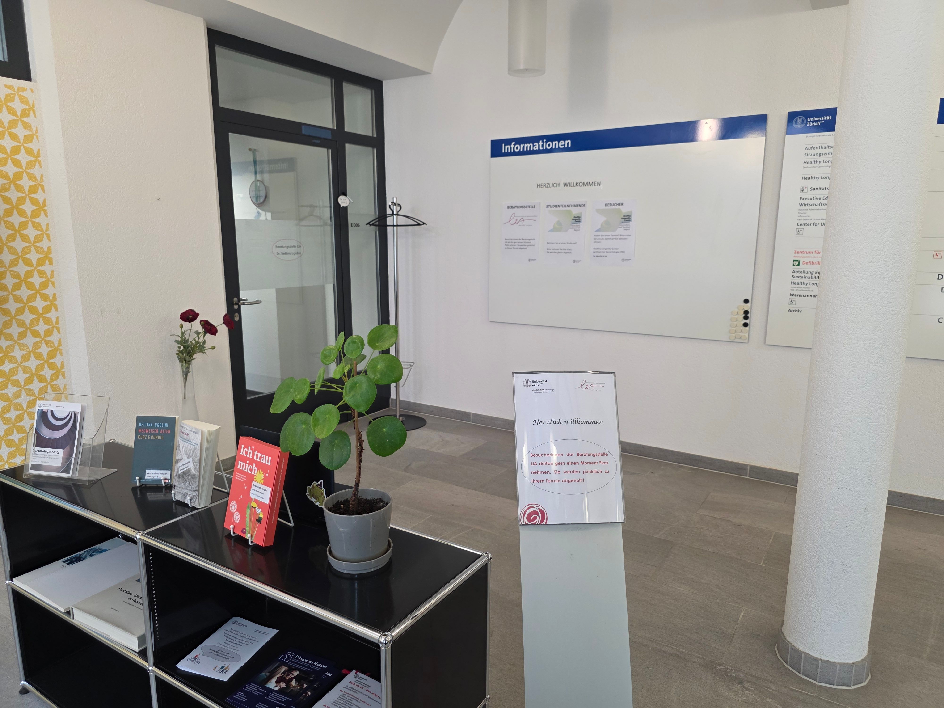 Foyer E-805: Eingangsbereich von der Beratungsstelle Leben im Alter. Rechts von der Tür befindet sich ein Garderobenständer mit mehreren Kleiderbügeln. In der Mitte vom Bild sieht man ein kleines Willkommensschild. Auf dem Schild steht, dass Besucher:innen einen Moment Platz nehmen dürfen, man wird für den Termin abgeholt. Links vom Schild hat es ein kleines schwarzes Regal mit verschiedenen Flyern.