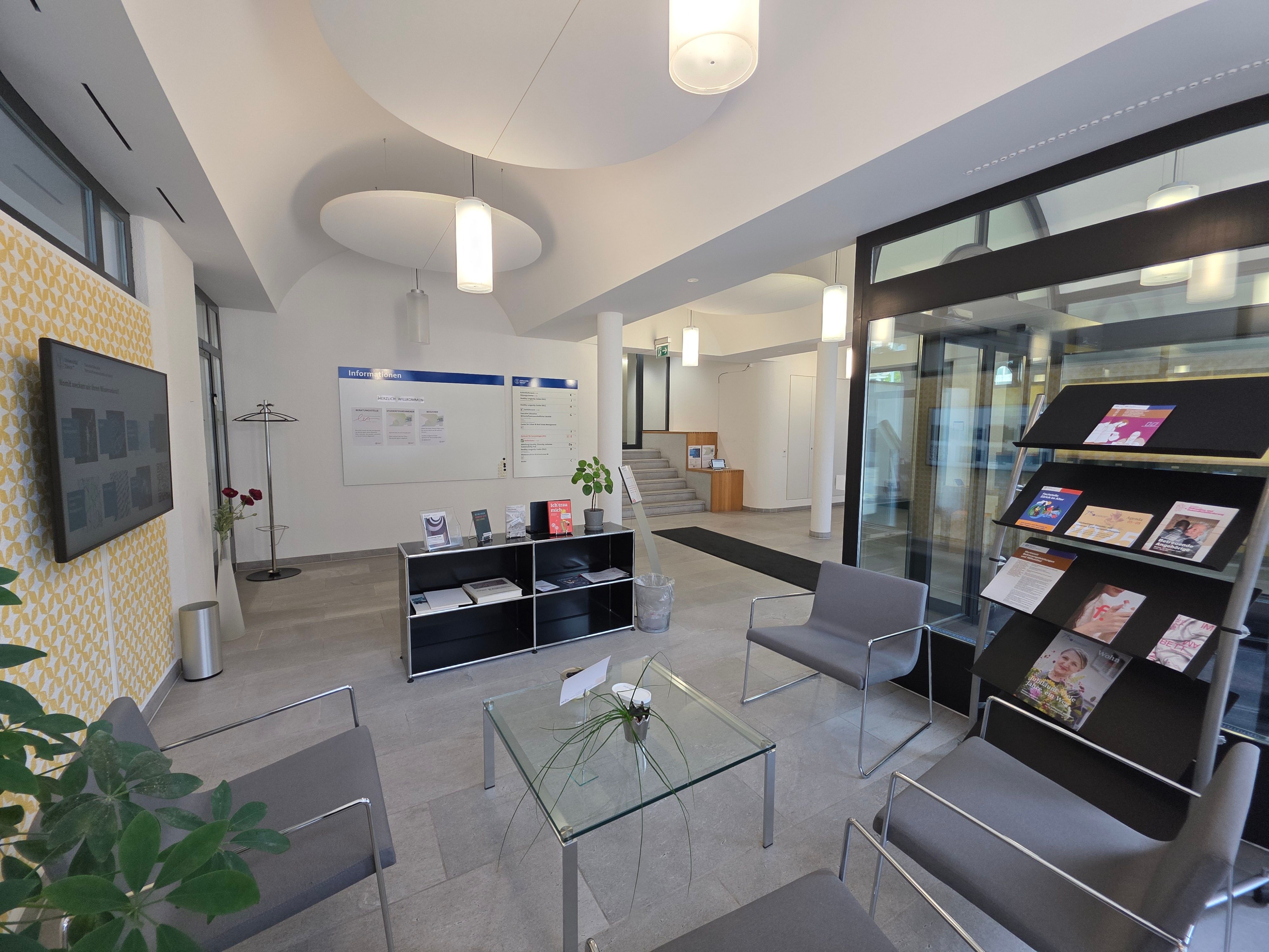 Foyer E-805: Wartebereich der Beratungsstelle im Alter. Blick auf das ganze Foyer vom Fenster aus. Vier graue Stühle stehen rund um einen viereckigen Glastisch. Daneben befinden sich zwei Regale mit Broschüren und Flyer zum Thema Alter.