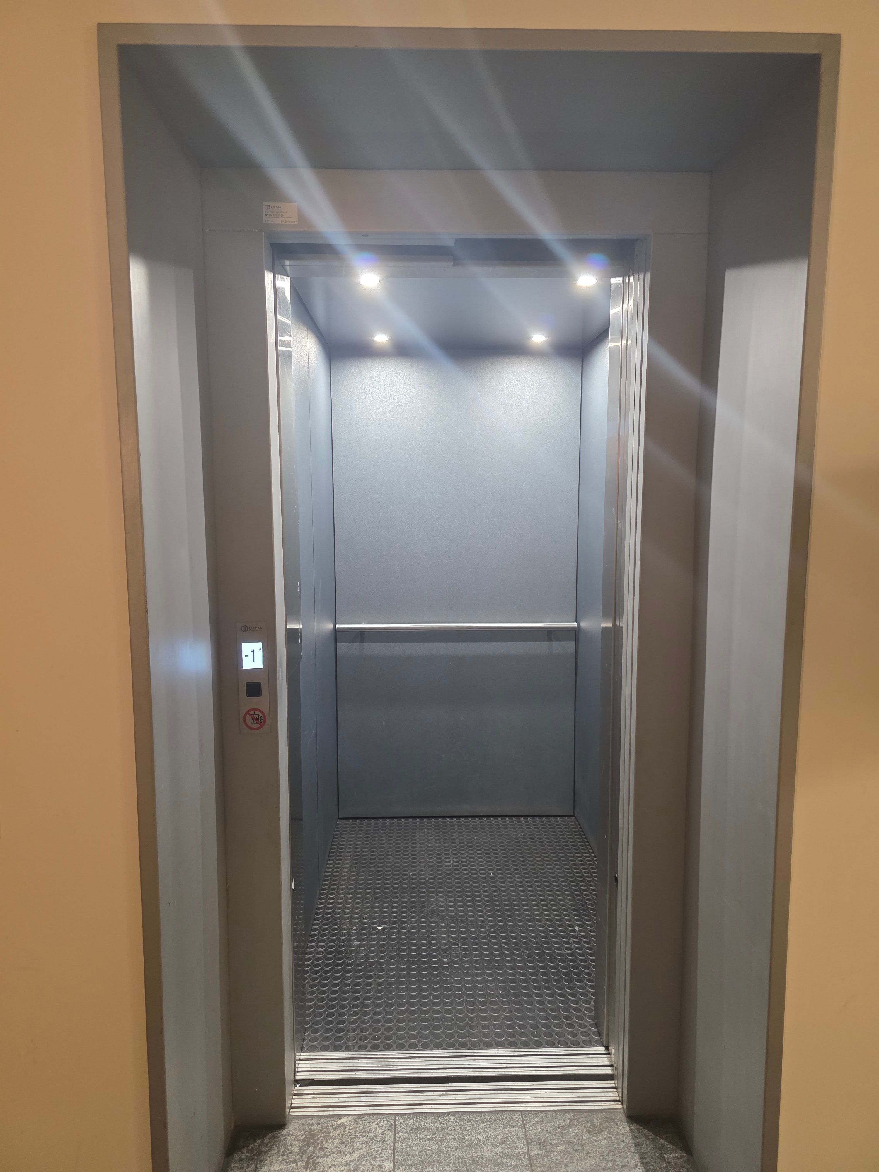 PET Lift: Bild von einem schmalen Lift. An der Aussenseite oberhalb der Lifttaste hat es ein Display. Darauf sieht man auf welchem Stockwerk der Lift ist. Die automatische Lifttüre ist geöffnet.