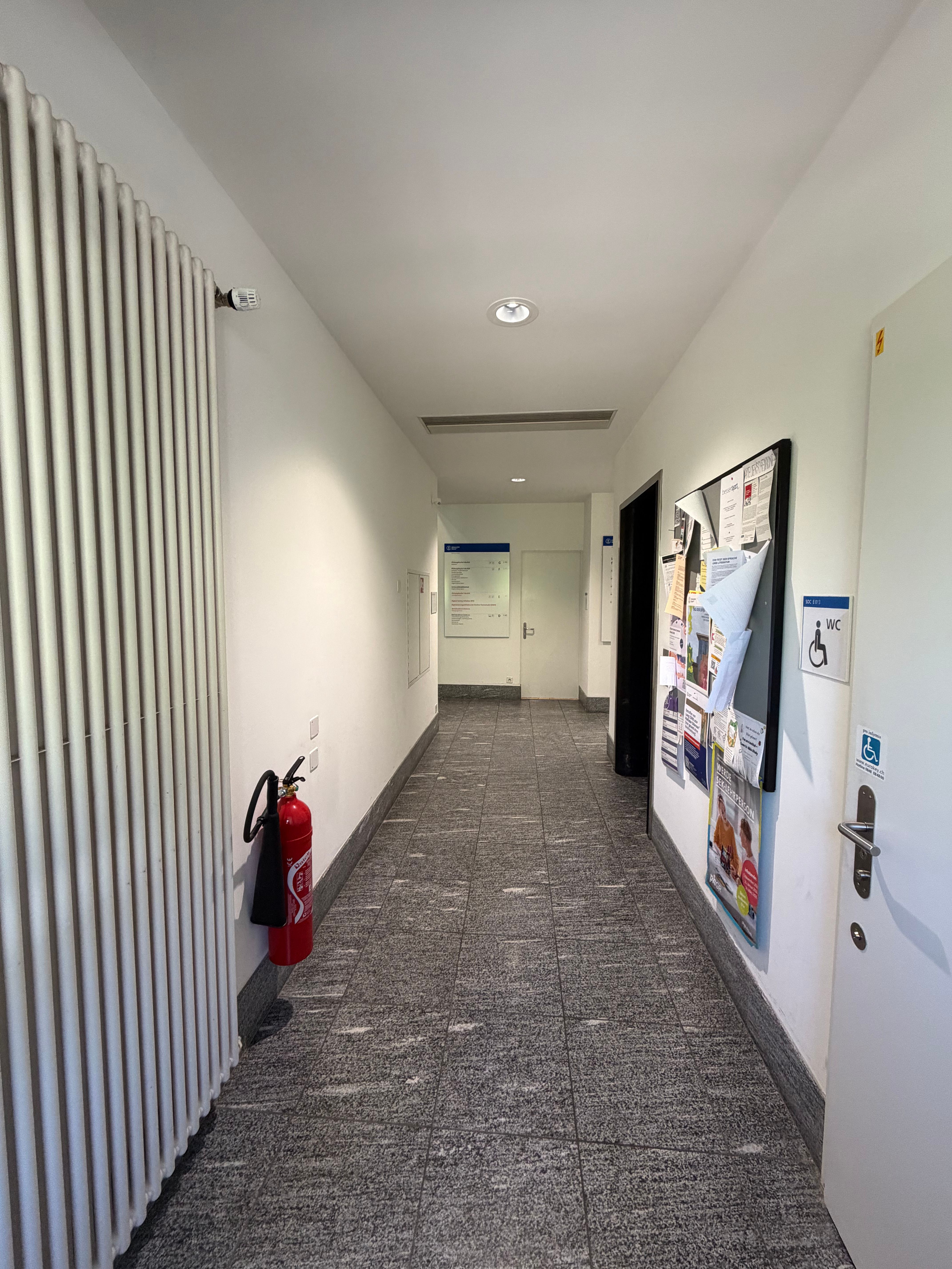 SOC_Stockwerk E (= EG):  Man sieht den Gang, in dem der Lift im EG vorhanden ist. Fotografiert vom Fenster aus mit Sicht auf die Herrentoilette am Ende des Gangs beim Übergang ins Treppenhaus. Der Lift ist auf der rechten Seite, davor das Rollstuhl-WC und Anschlagebrett. Links ist ein grosser Heizkörper an der Wand festgemacht, sowie der Feuerlöscher. Rechts geht der Weg zum Büro "Reinigungsdienst" des Betriebsdienstes Zentrum (Raum SOC E-012), links geht der Weg zum Treppenhaus des Erdgeschosses (Stockwerk E).