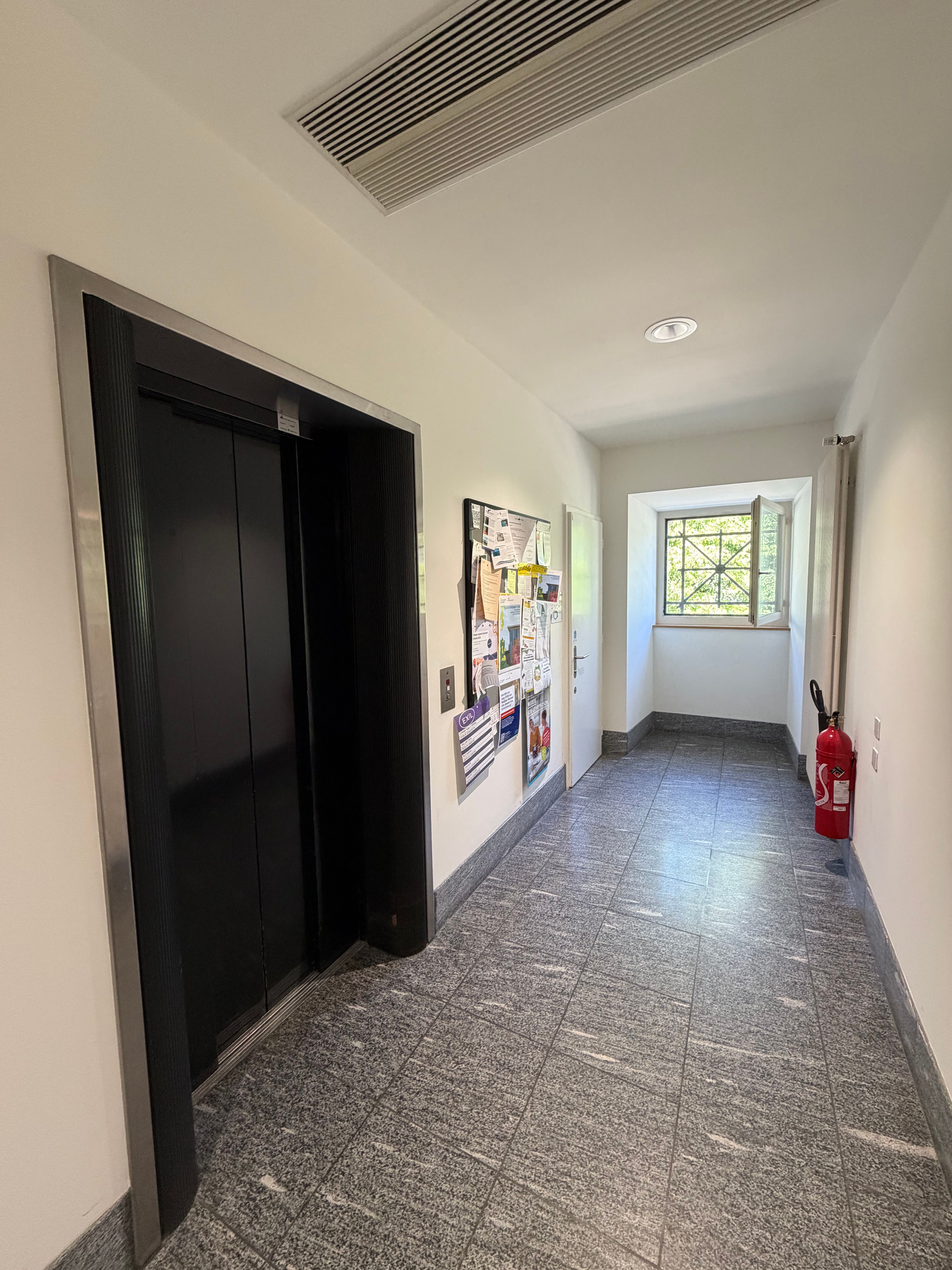 SOC_Stockwerk E (= EG):  Man sieht den Gang, in dem der Lift im EG vorhanden ist. Sicht auf das Fenster am Ende des Gangs. Der Lift ist auf der linken Seite. Dahinter ist ein Anschlagbrett und dann folgt das Rollstuhl-WC (mit Eurokey). Rechts ist ein grosser Heizungskörper an der Wand, sowie ein Feuerlöscher.
