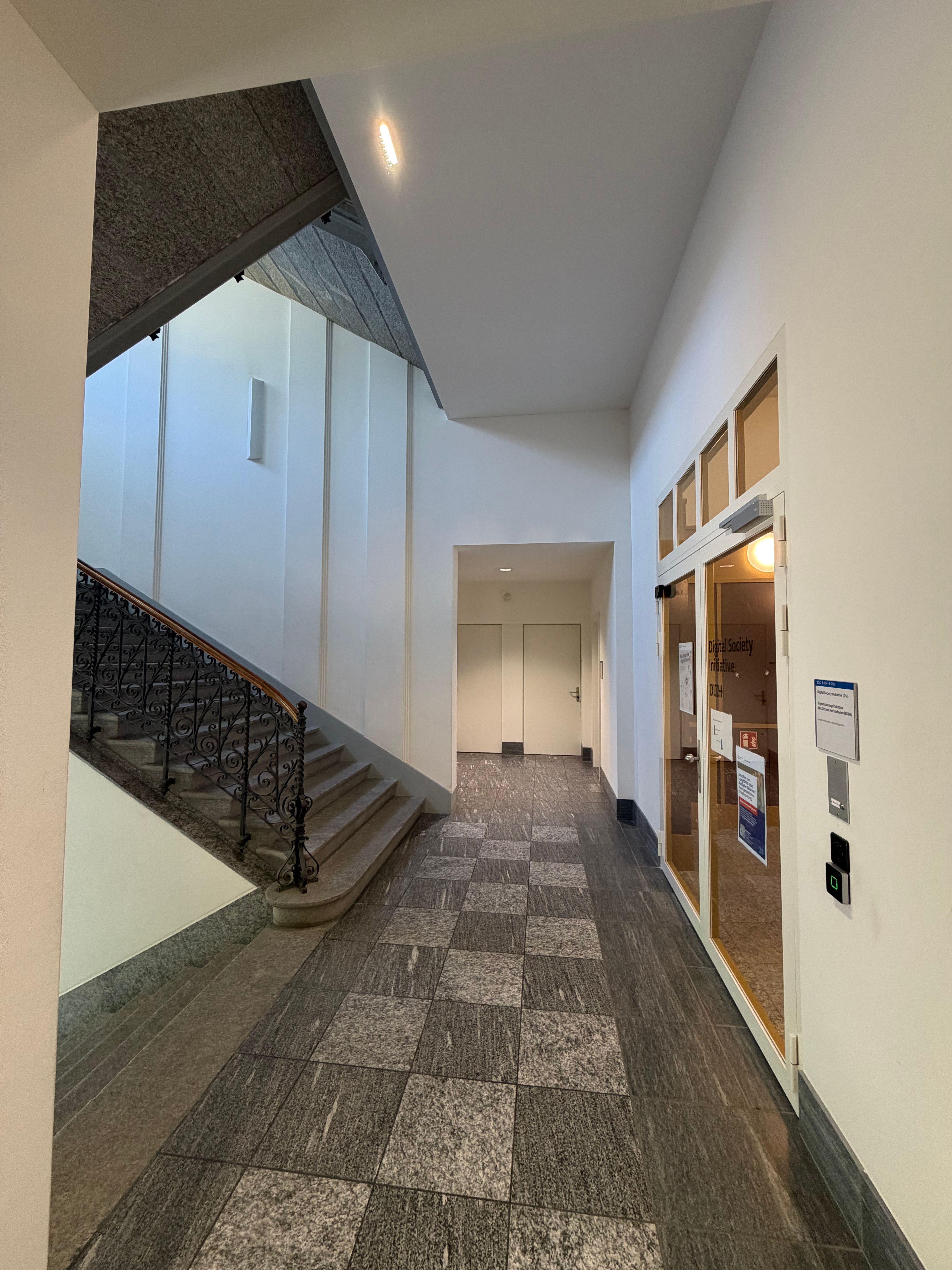 SOC_Stockwerk E (= EG): Treppenhaus des Erdgeschosses (Stockwerk E) mit Blick in Richtung Sitzungszimmer und Gruppenräume (SOC E-002). Auf der linken Seite werden die Treppen angedeutet, rechts führt der Weg durch die gesperrte Glastür zu den Räumlichkeiten der Digital Initiative Society.