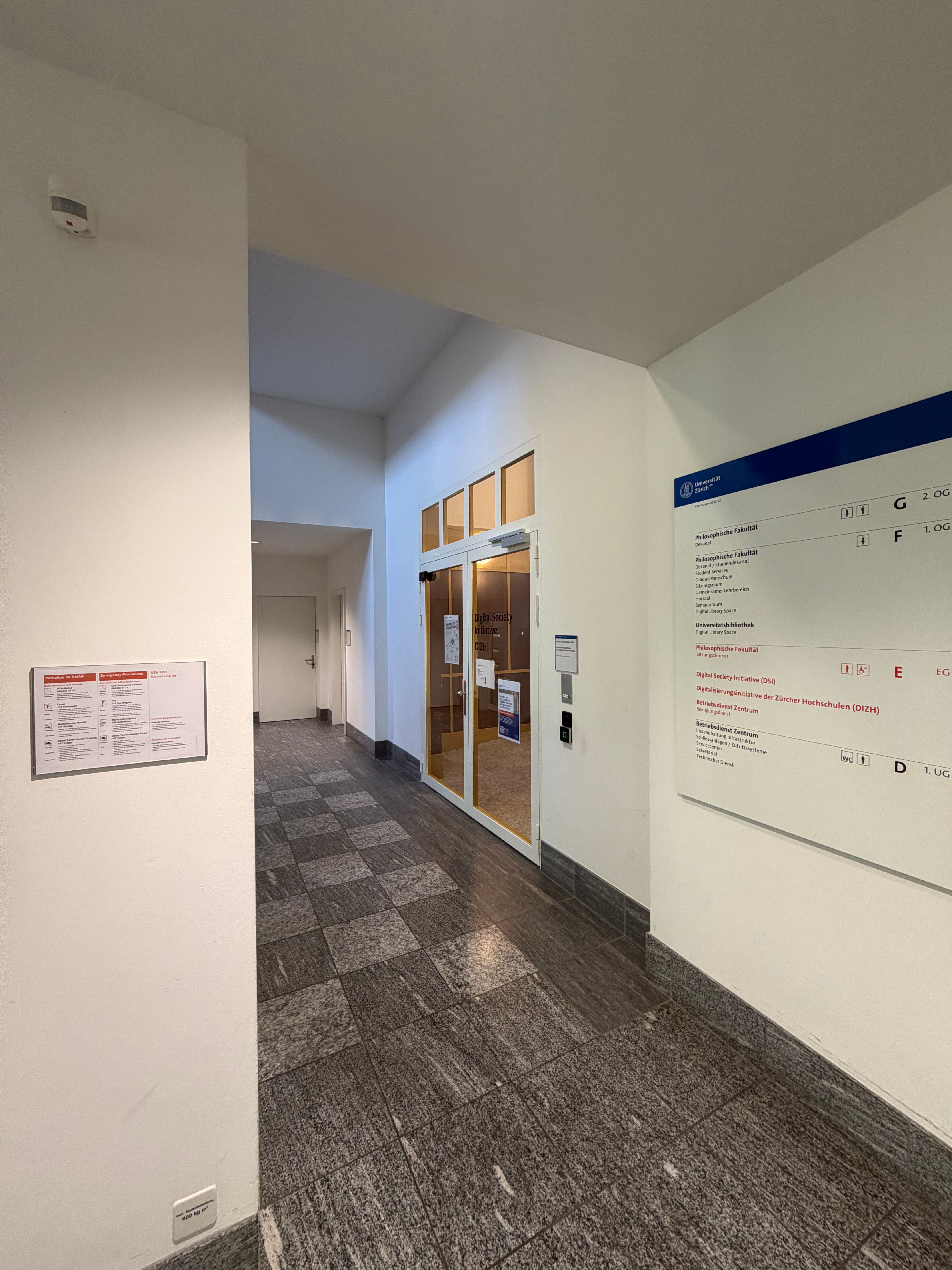 SOC_Stockwerk E (= EG): Blick vom Lift aus in Richtung Treppenhaus und Räumlichkeiten des Stockes E. Auf der rechten Seite ist die Übersichtstafel des Stockwerkes abgebildet, dahinter die Glastüre, welche den Zugang zur Digital Initiative Society der Universität Zürich verspricht. Direkt gegenüber werden die Sitzungszimmer und Gruppenräume des Stockwerkes E angedeutet.