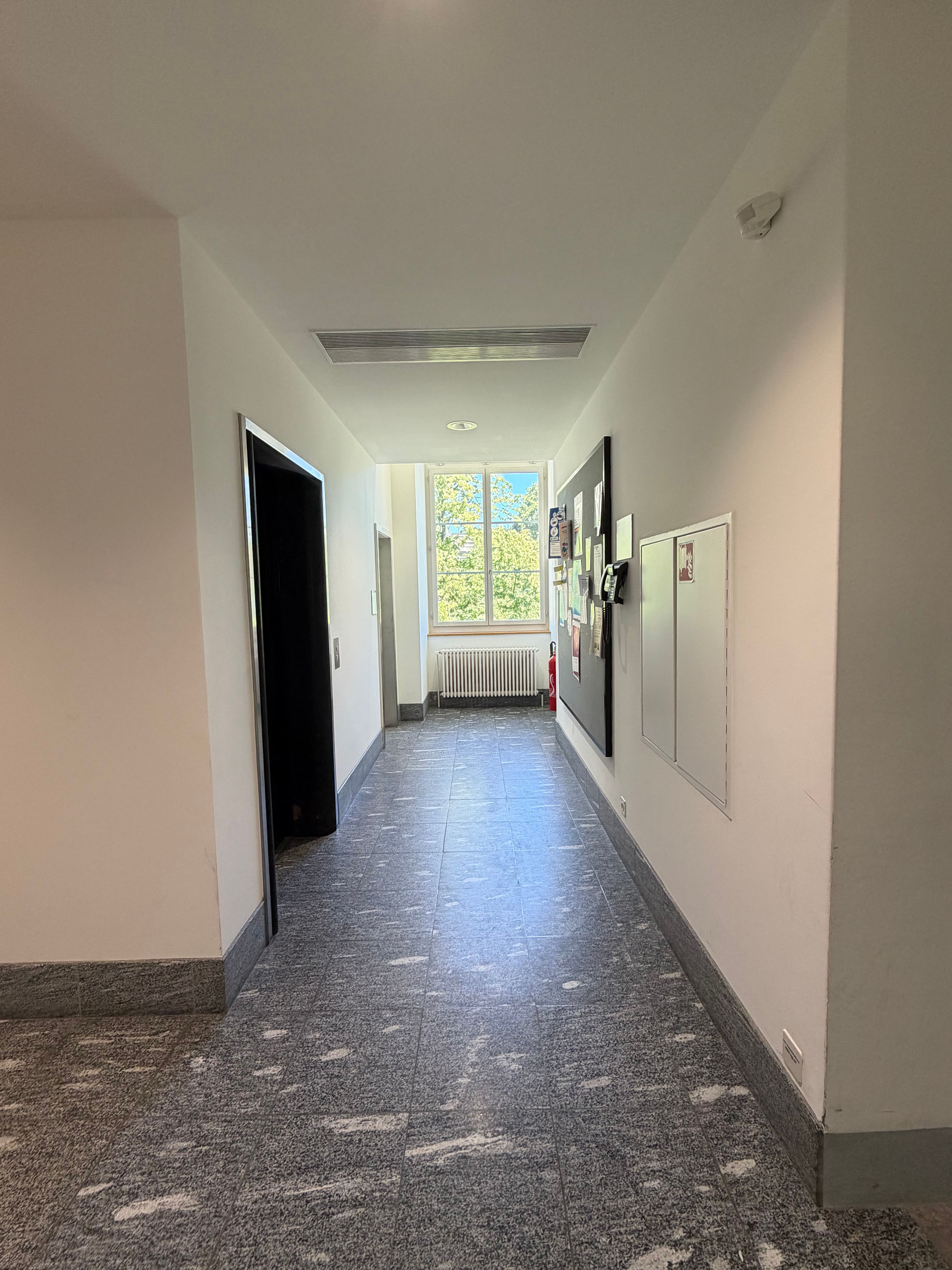 SOC_Stockwerk F (= 1. OG):  Man sieht den Gang, in dem der Lift im ersten OG vorhanden ist. Sicht auf das Fenster am Ende des Gangs. Der Lift ist auf der linken Seite, dahinter ein unbeschrifteter Raum. Rechts ist ein Anschlagbrett an der Wand aufgehängt, sowie Feuerlöscher und Brandschutzschrank.