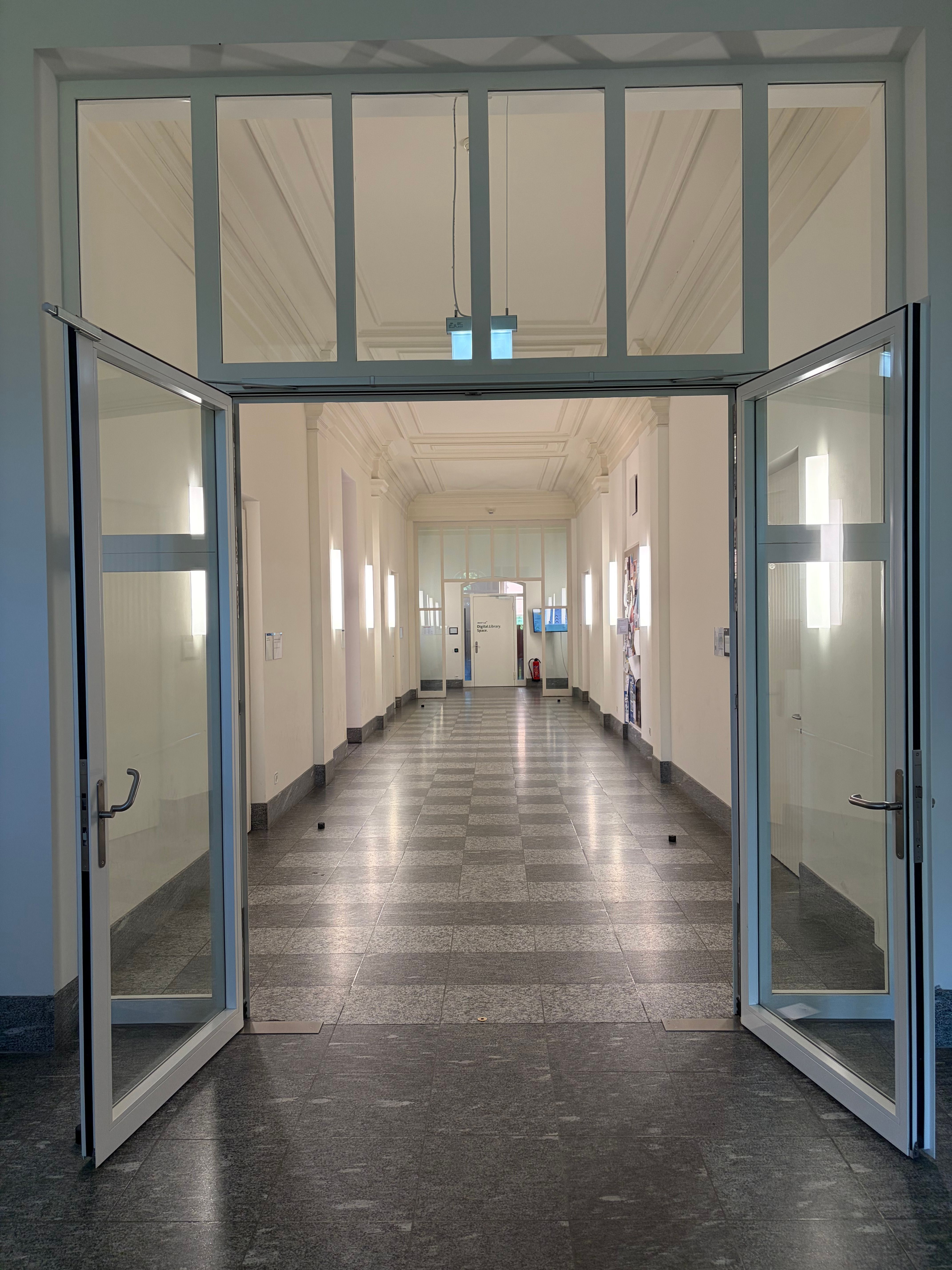 SOC_Stockwerk F (= 1. OG): Gang zu den Räumlichkeiten des Stockwerkes F (1. Obergeschoss). Fotografiert aus Sicht des Treppenhauses. Der Weg führt durch die offene Doppelglastür. Links befindet sich der grosse Hörsaal (SOC-F-106). Rechts befinden sich weitere Räumlichkeiten, von vorne nach hinten: SOC-F-102 "Geschaftsstelle und Service Desk Graduiertenschule", SOC-F-103 "Aufenthaltsraum mit Kopierer", SOC-F-104 "Sitzungsraum". Am Ende des Ganges befindet sich die Universitätsbibliothek "Digital Library Space". Am Boden sind die Boden-Türstopper mit Arretierung zu sehen.