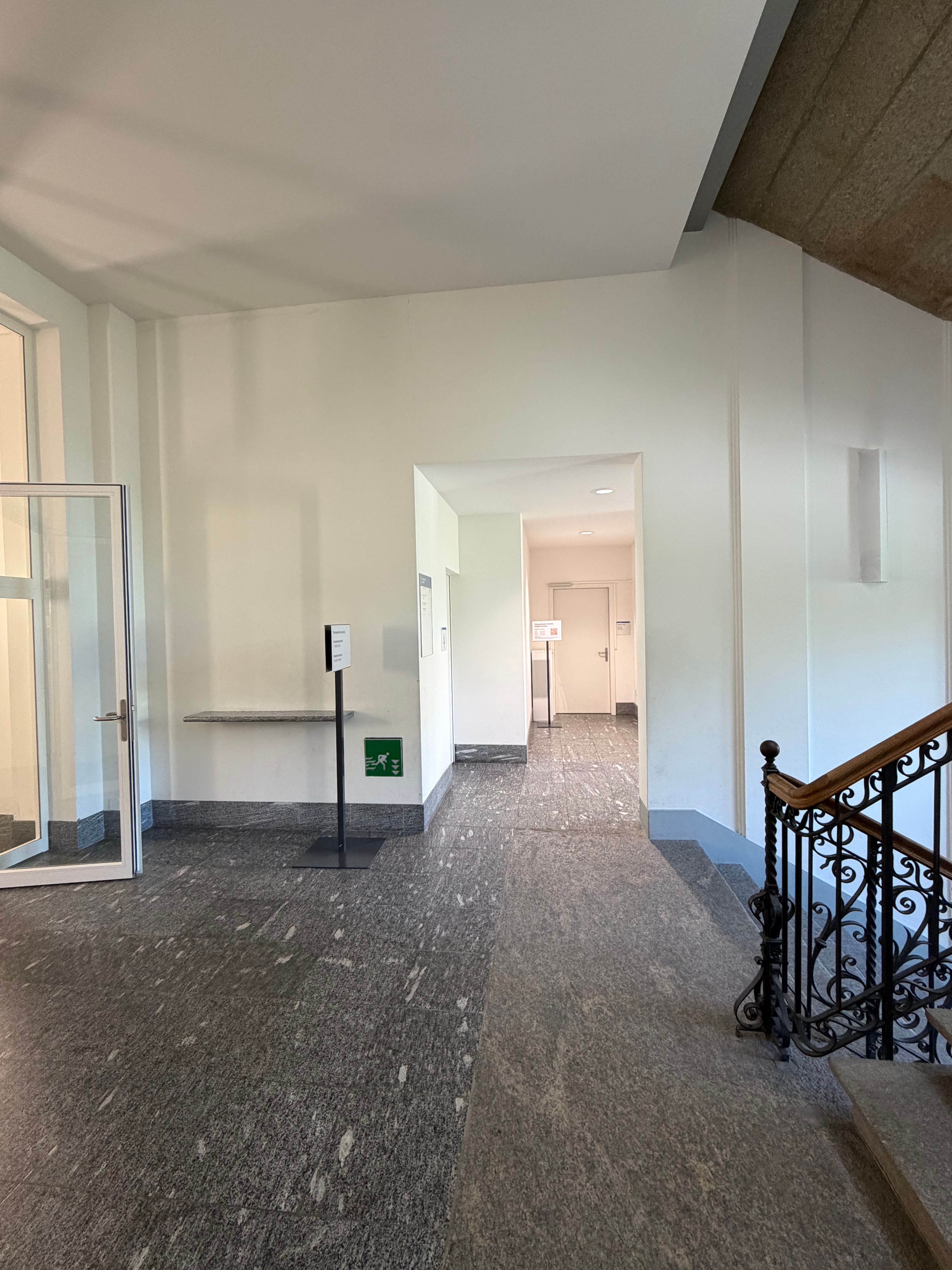 SOC_Stockwerk F (= 1. OG): Treppenhaus des ersten Obergeschosses (Stockwerk F) mit Blick in Richtung Büro "Student Services" (SOC-F-108b) der philosophischen Fakultät. Auf der rechten Seite werden die Treppen angedeutet, links führ der Weg durch die offene Glastür zu den Räumlichkeiten des Stockwerkes.