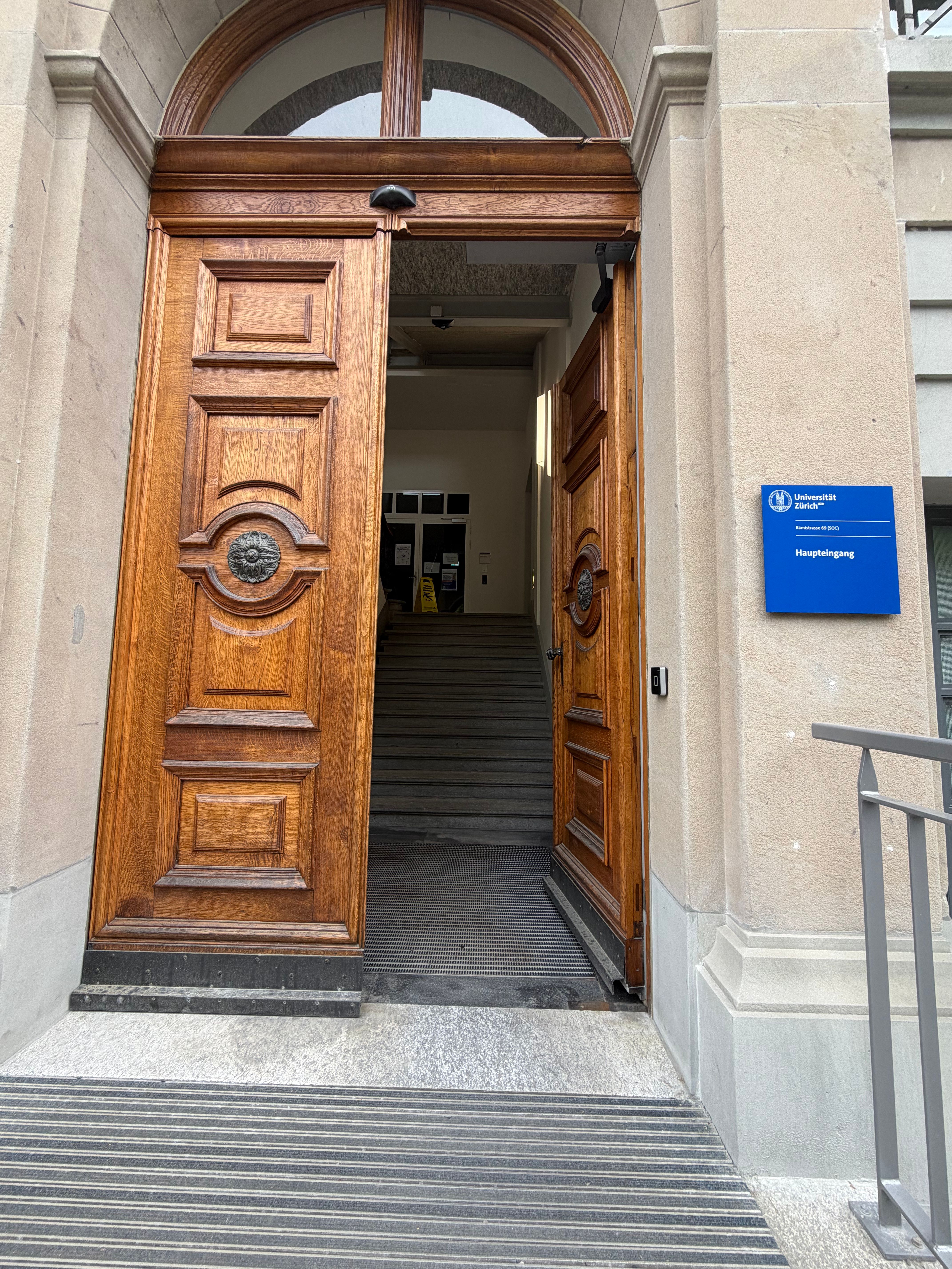 Offene Türe des Haupteinganges. Der Haupteingang ist nur über Treppe erreichbar. Beim Eintreten des Gebäudes folgt noch eine Treppe hoch zu Stockwerk E (= EG). Für einen stufenlosen Zugang zum Gebäude gibt rechts neben dem Haupteingang den Zugang zum Lift.