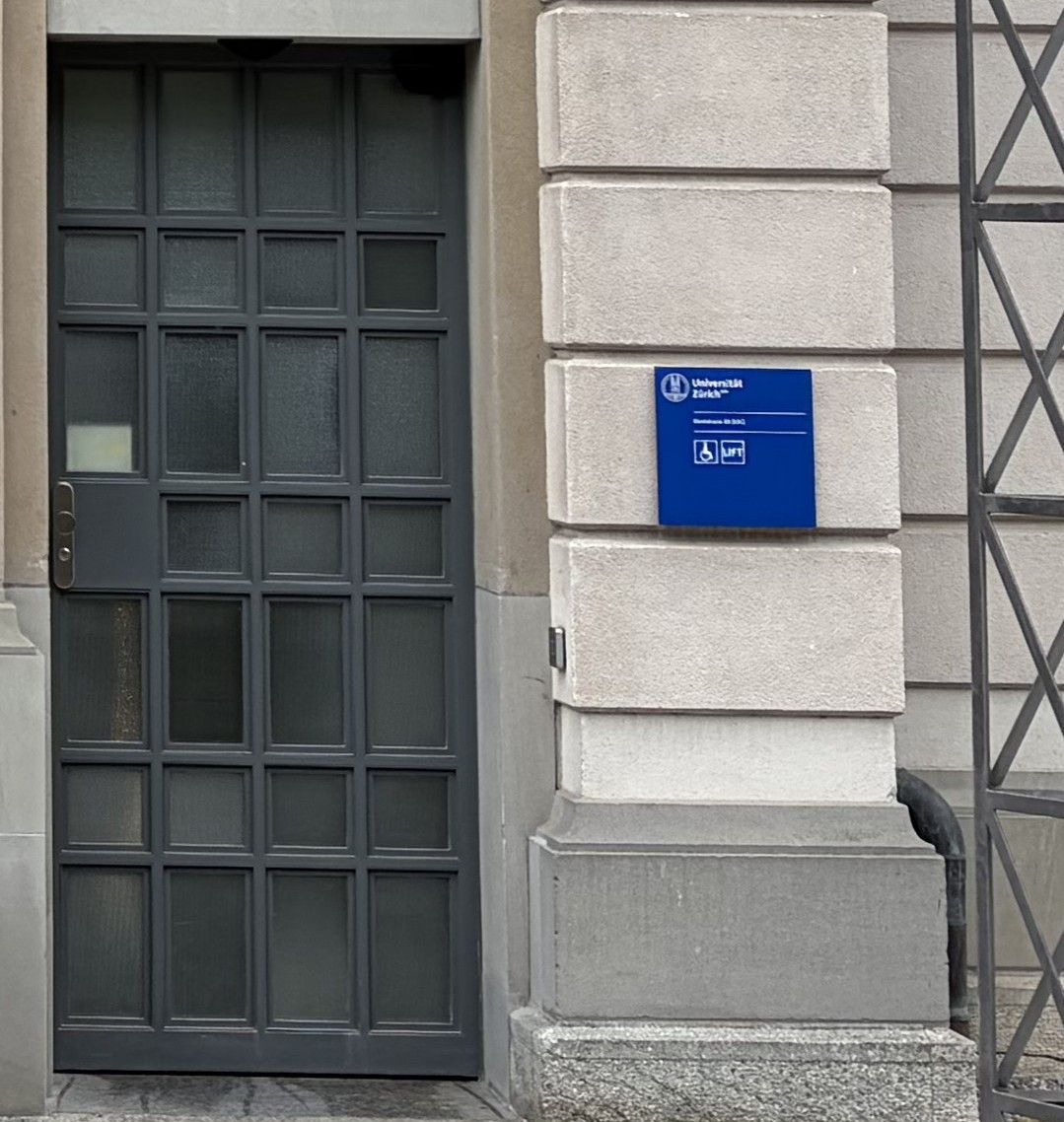Gebäude SOC: Abgebildet ist der stufenlose Eingang von aussen. Die Tür ist geschlossen und öffnet sich automatisch gegen innen. Auf der rechten Seite ist die Signaletik für Rollstuhl und Lift angebracht.