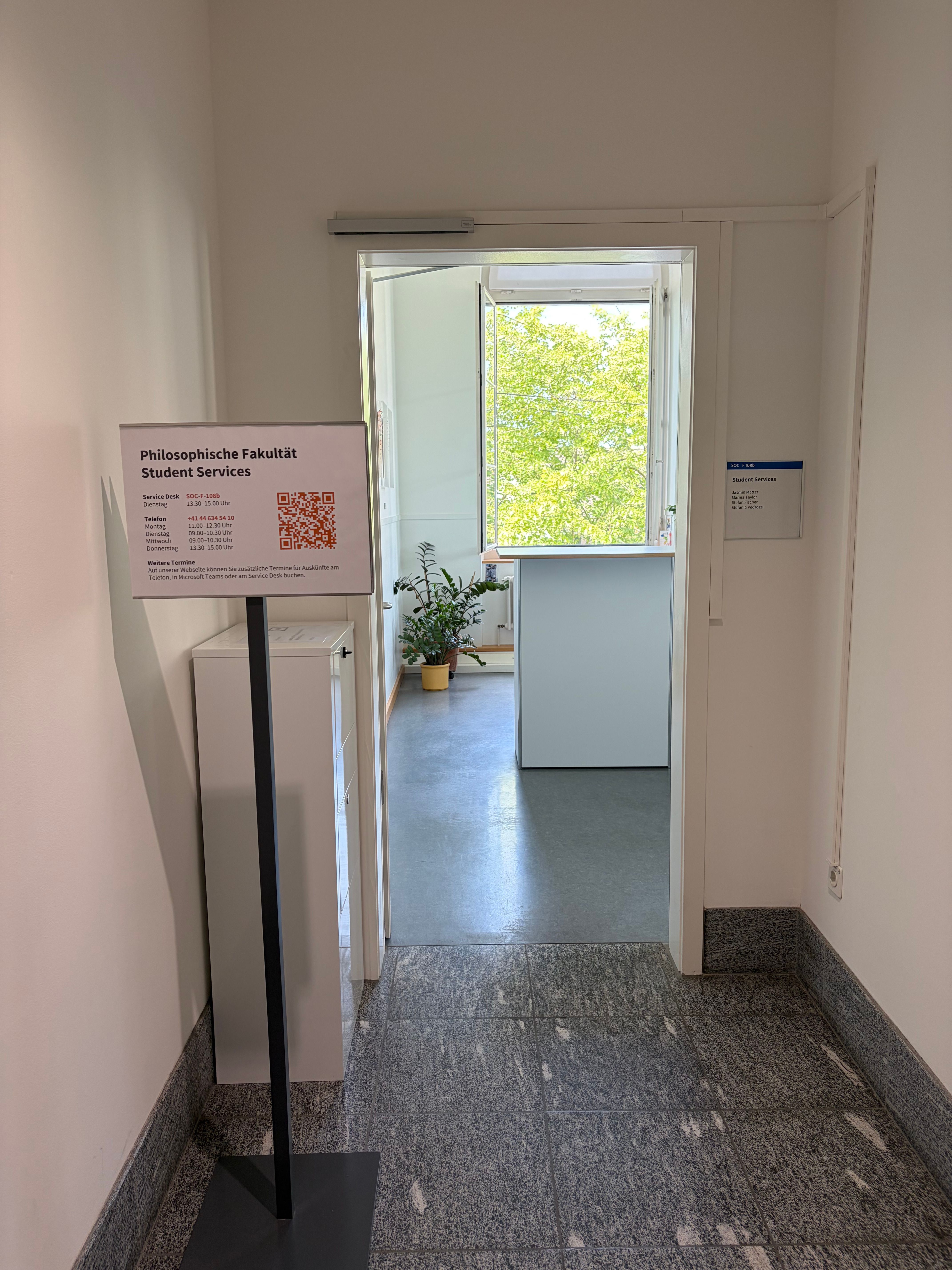 SOC F-108: Abgebildet ist der Eingang zum Service Desk der Philosophischen Fakultät. Die Türe ist offen und es ist der Empfangstresen innerhalt des Raumes zu sehen. 
Vor der Tür links steht eine Informationstafel mit den Informationen zu den Öffnungszeiten. Dahinter steht ein Korpus. Es handelt sich dabei um den Briefkasten.