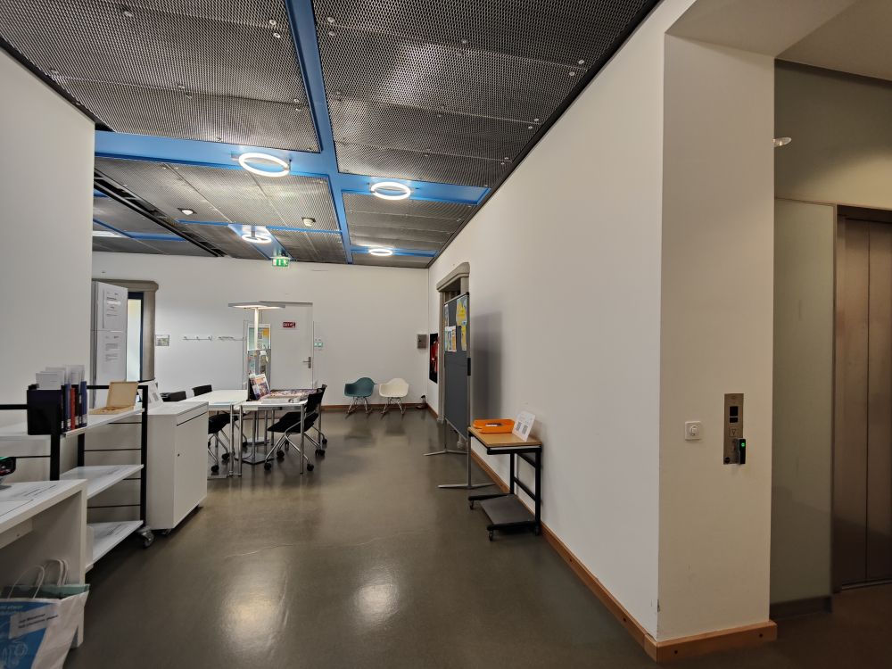 Arbeitsraum für Studierende KOL-H-303c: Foyer vom Lesesaal aus gesehen. Rechts befindet sich der Lift Ost. Links befinden sich die Selbstausleihe, der Bereitstellungswagen und der Rueckgabekasten. Hinten in der Mitte befindet sich die Eingangstuer und rechts davon der Kopierraum.