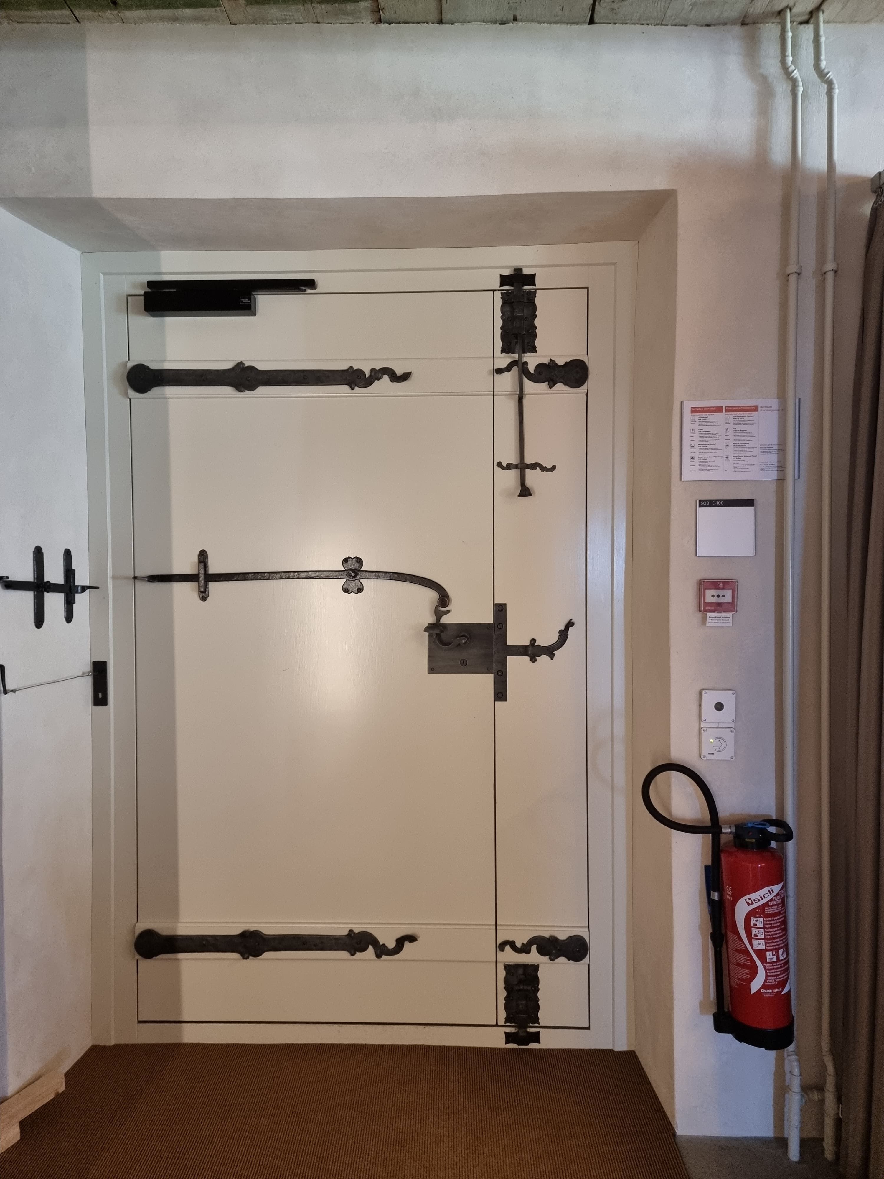 SOB Gebäude Erdgeschoss: Auf dem Bild ist der Hauptausgang des SOB Gebäudes abgebildet. Rechts des Ausgangs befindet sich eine der Brandmeldeanlagen, dessen Scheibe man nicht einschlagen muss, und ein Feuerlöscher.