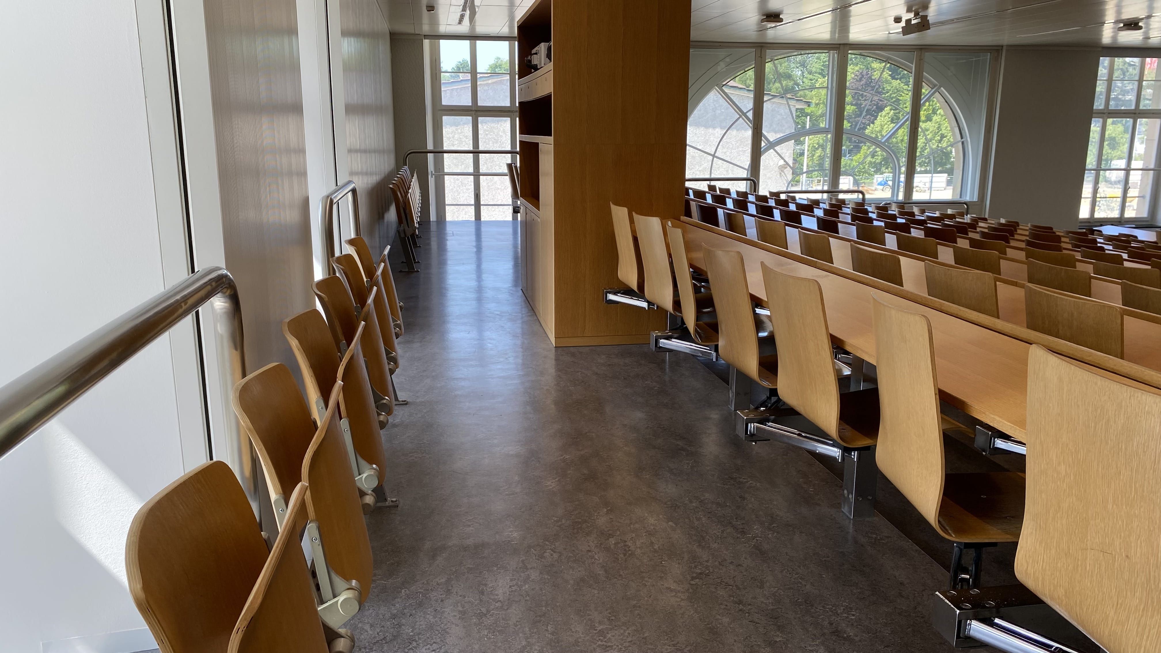 Hörsaal RAI-H-041: Ansicht auf die hinterste Reihe. Der Durchgang zwischen den Sitzmöglichkeiten mit und ohne Tisch ist breit. Auf dieser Ebene gibt es ebenfalls eine Eingangstüre.