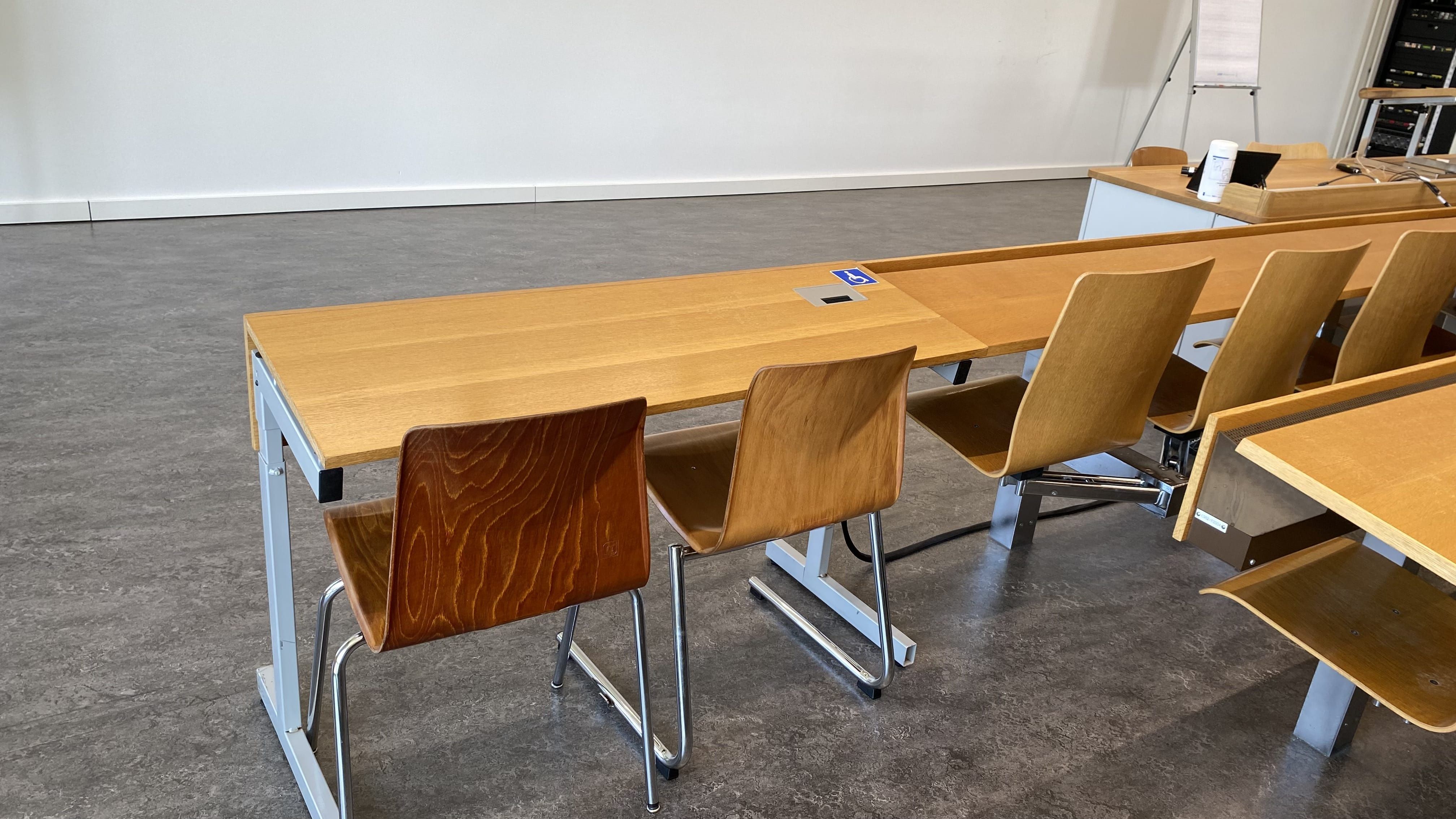 Hörsaal RAI-H-041: Ansicht auf den zweiten Tisch mit Rollstuhl-Signet in der vordersten Reihe ohne fixe Bestuhlung. Dieser Tisch befindet sich auf der linken Seite des Raumes.