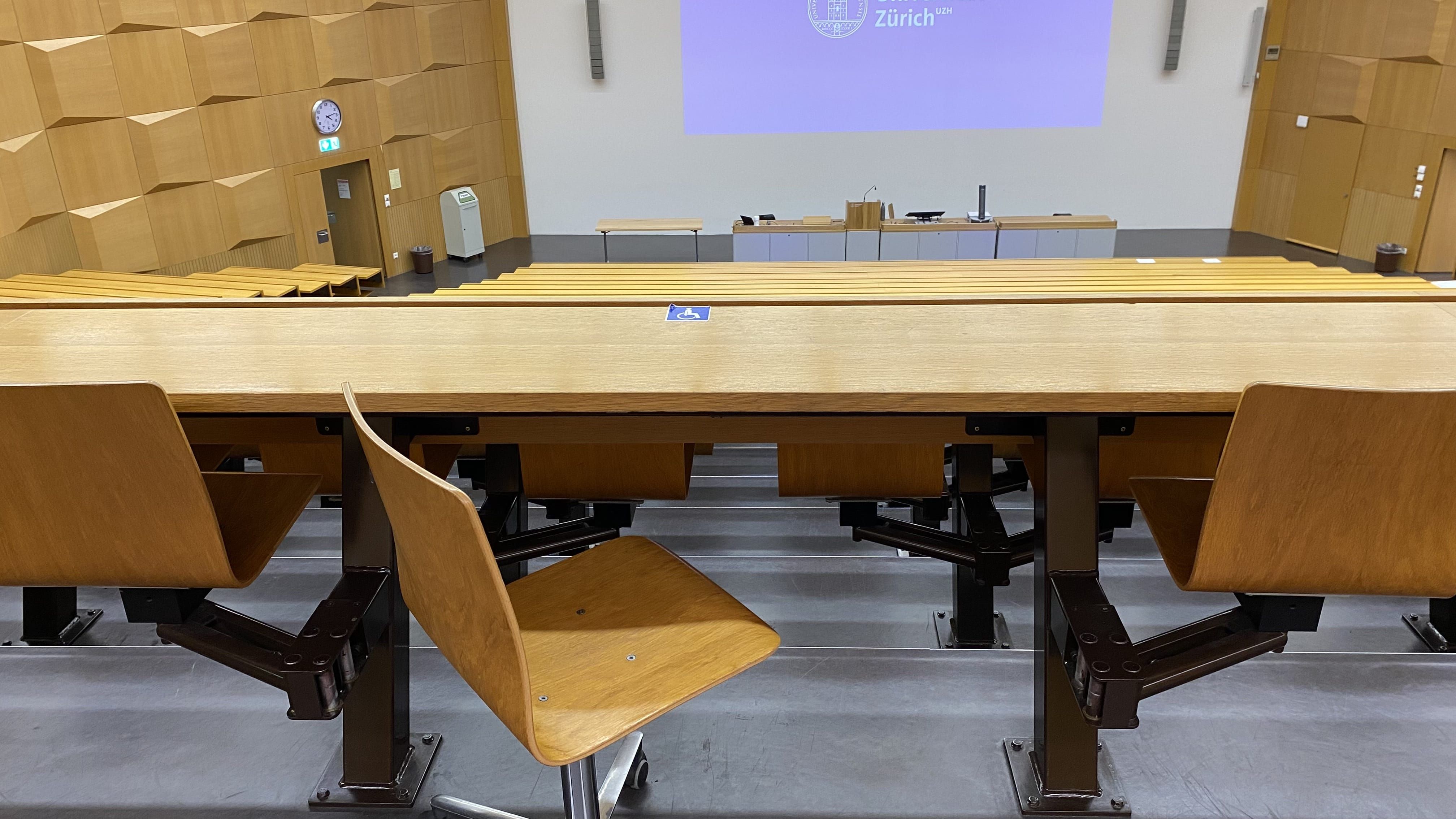 Hörsaal HAH-E-03: Ansicht auf einen Tisch mit Rollstuhl-Signet, ohne fixe Bestuhlung. Der Tisch befindet sich in der obersten Reihe in der Mitte, und ist stufenlos durch die Eingangstüren oben links oder rechts erreichbar.