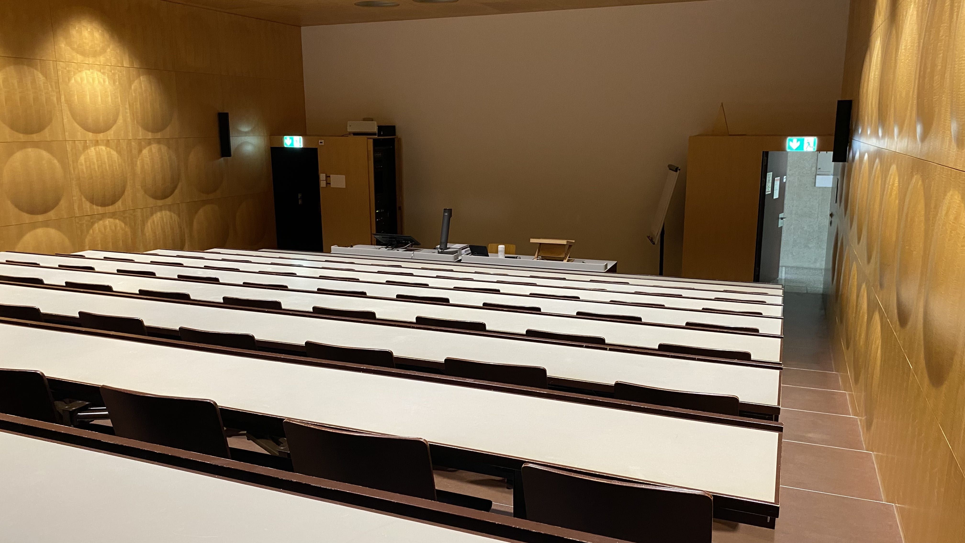 Hörsaal HAH-F-01: Blick aus der obersten Reihe zum Dozierendenpult hin. Links und rechts sind beide Eingangstüren ersichtlich.