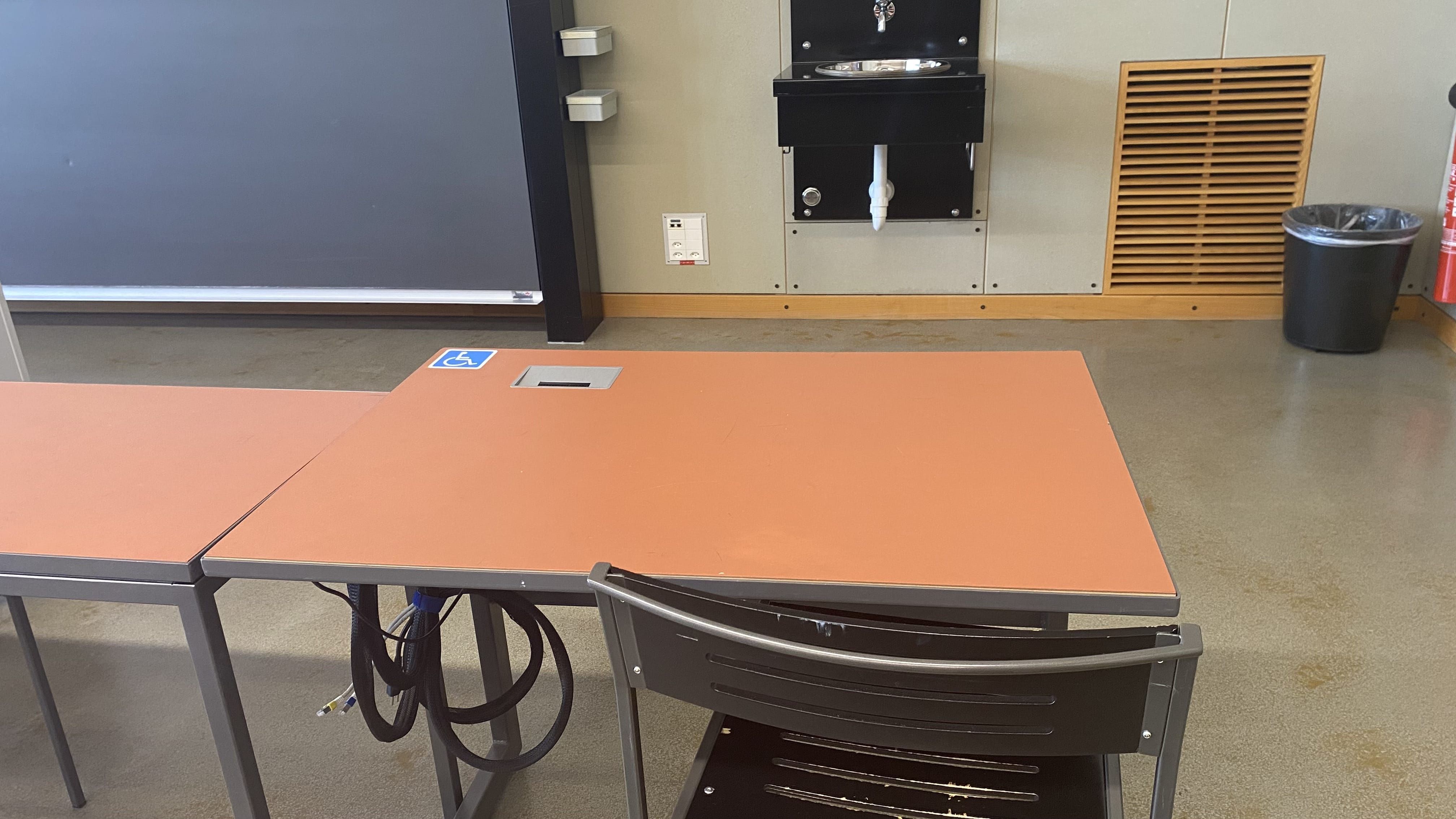 Hörsaal KOL-E-18: Ansicht auf den Tisch mit Rollstuhl-Signet, elektrisch verstellbar. Der Tisch befindet sich in der ersten Reihe auf der rechten Seite. Der Tisch ist nahe der Eingangstür auf der Seite des Dozierendenpults.
