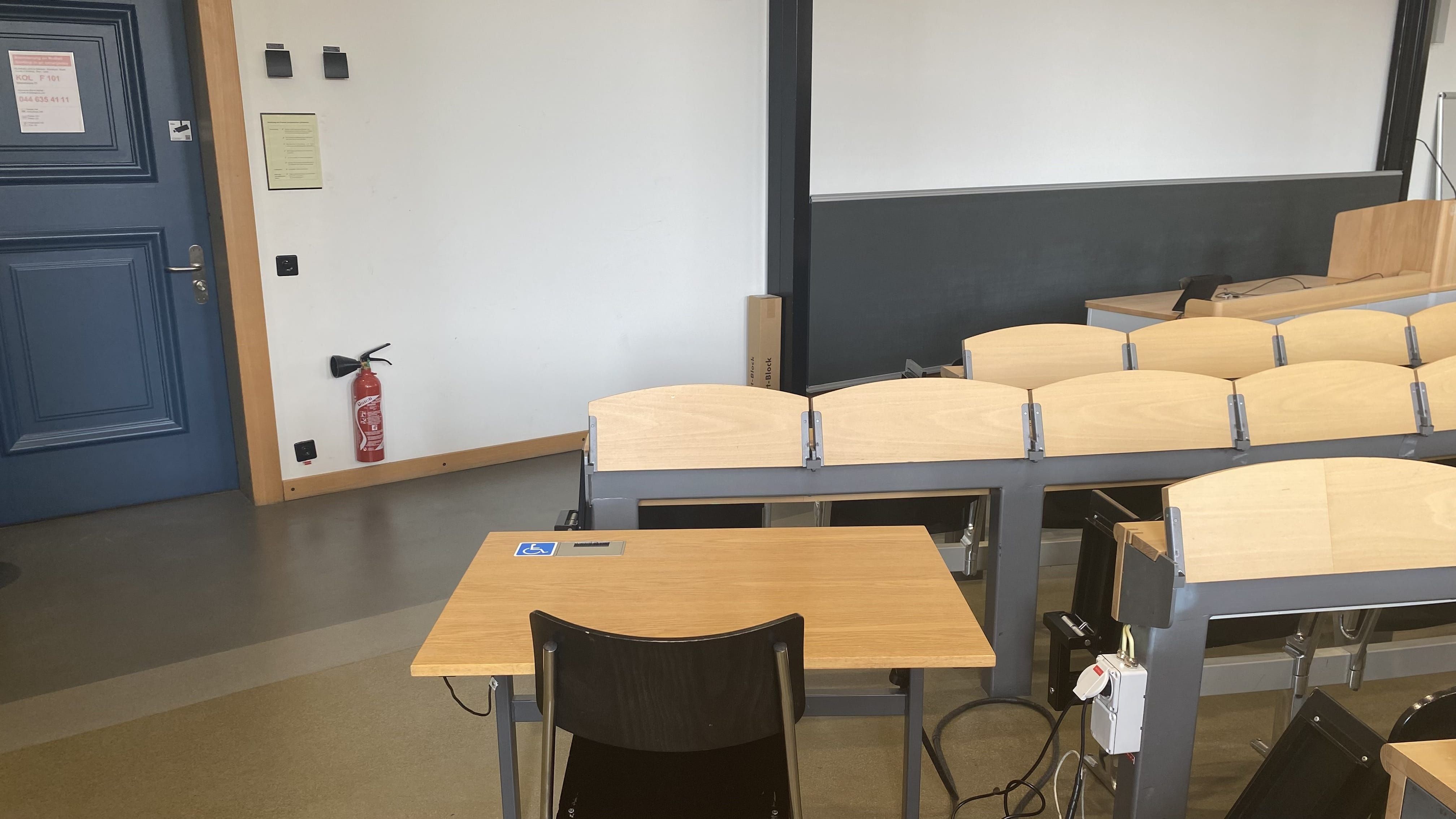 Hörsaal KOL-F-101: Ansicht auf den elektrisch höhenverstellbarer Tisch mit Rollstuhl-Signet. Der Tisch befindet sich in der dritten Reihe nahe der rechten Eingangstür.
