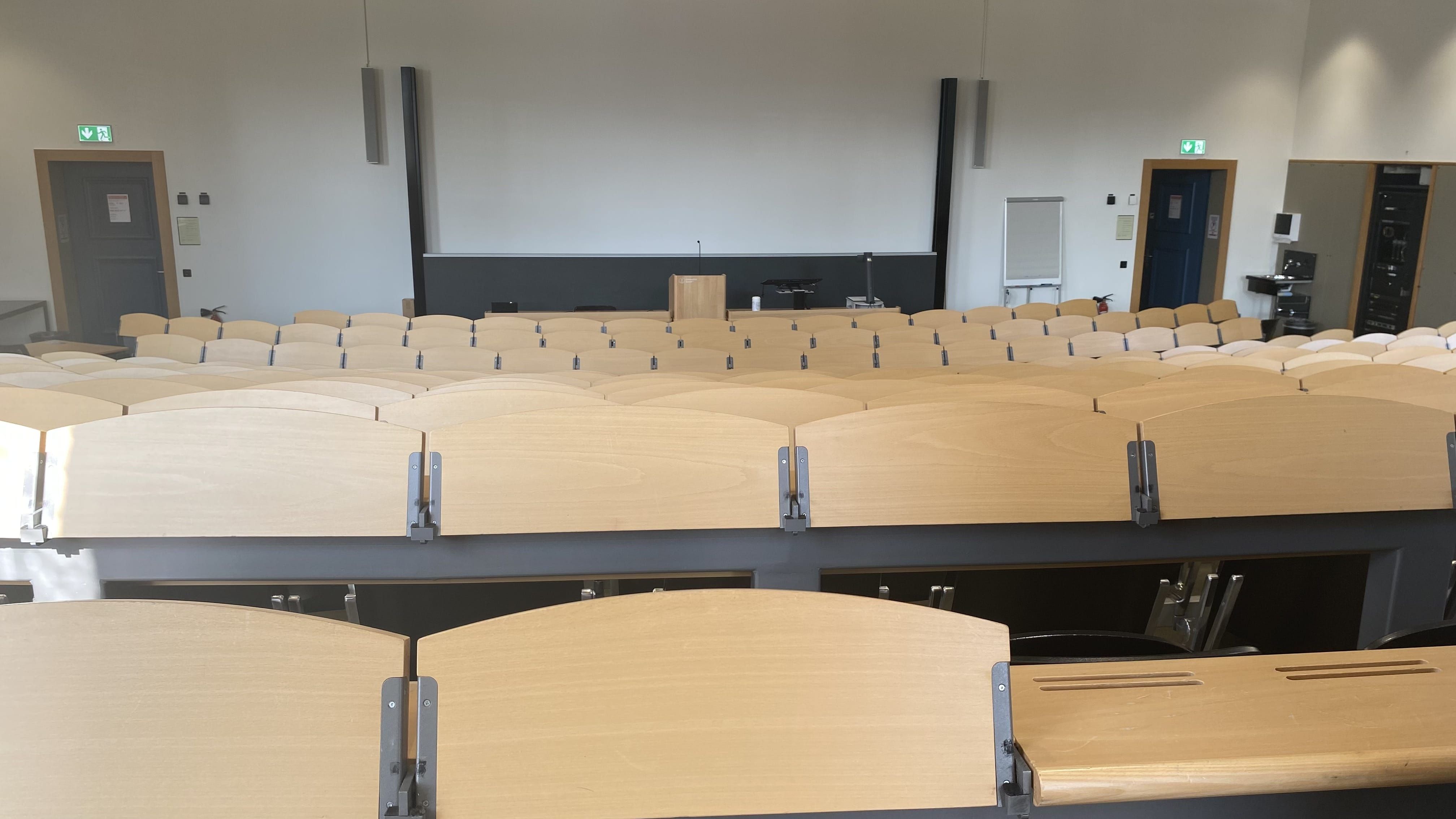 Hörsaal KOL-F-101: Ansicht zum Dozierendenpult hin. Die Eingangstüren befinden sich links und rechts im Bild. Neben der rechten Eingangstüre befindet sich ein Lavabo.