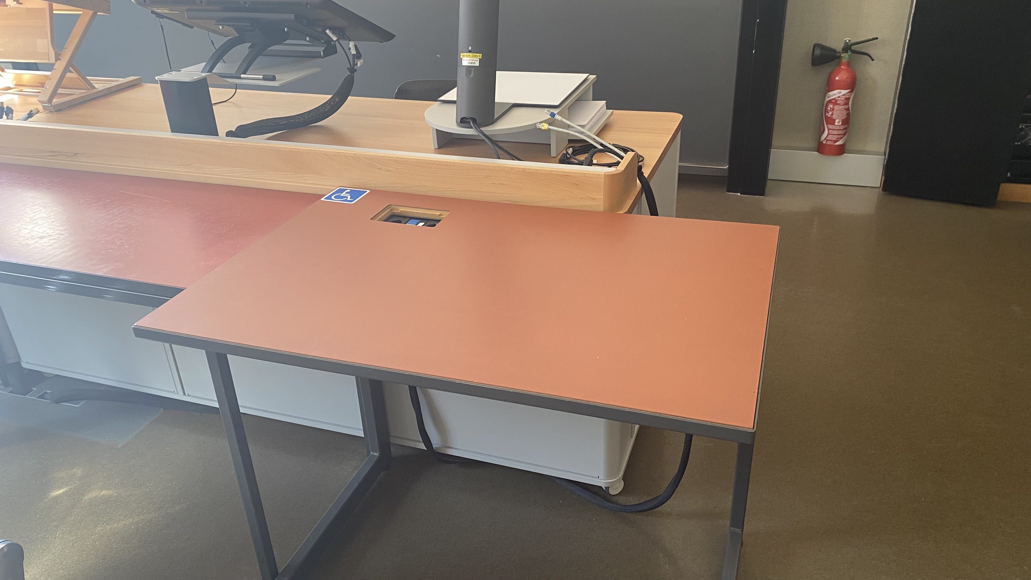 Hörsaal KOL-E-21: Ansicht auf den elektronisch verstellbaren Tisch mit Rollstuhl-Signet. Der Tisch befindet sich in der vordersten Reihe gleich vor dem Dozierendenpult.