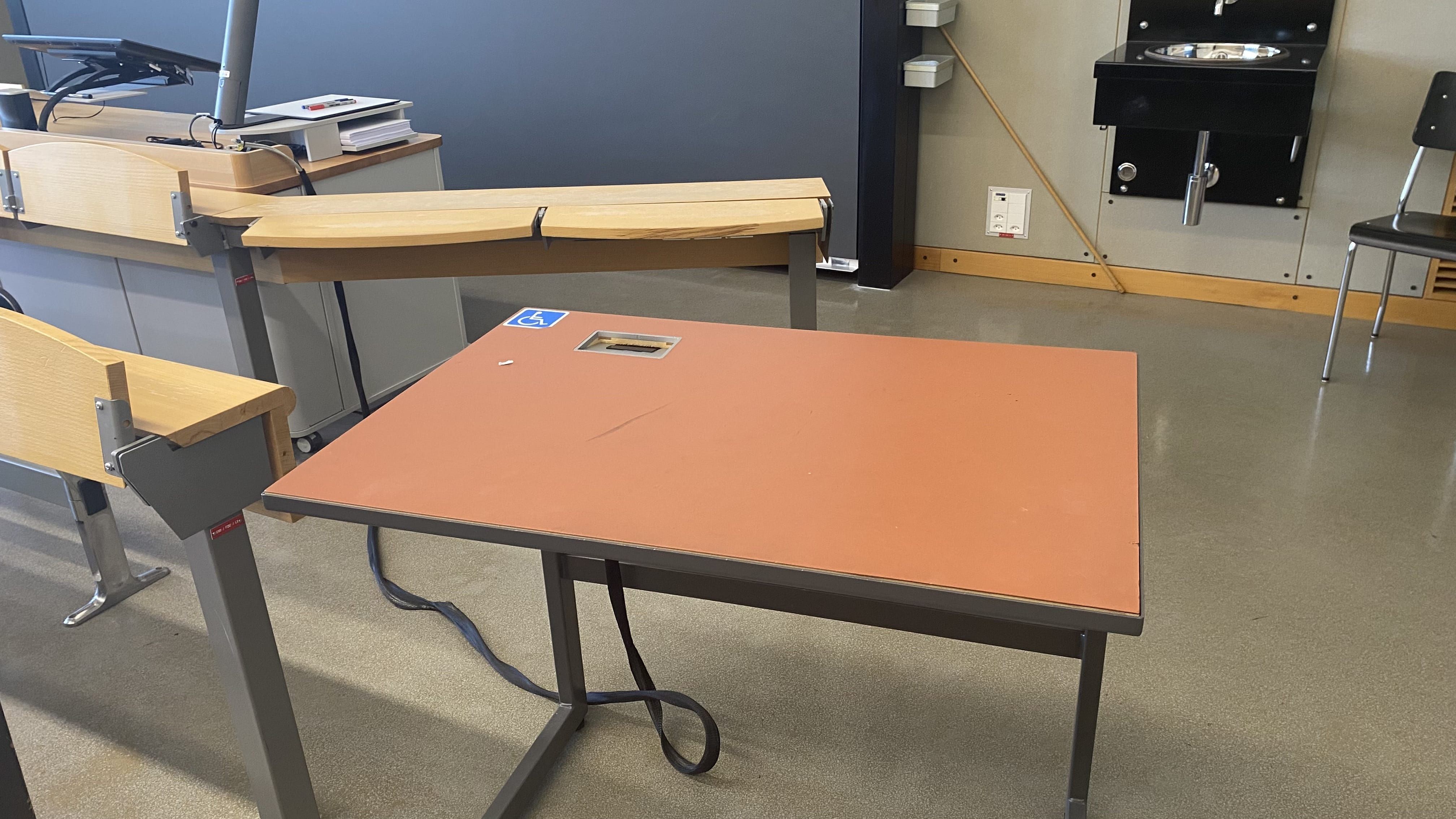Hörsaal KOL-F-117: Ansicht auf den elektrisch höhenverstellbaren Tisch mit Rollstuhl-Signet auf der rechten Seite des Raumes. Der Tisch befindet sich in der zweiten Reihe, nahe der rechten Eingangstür.