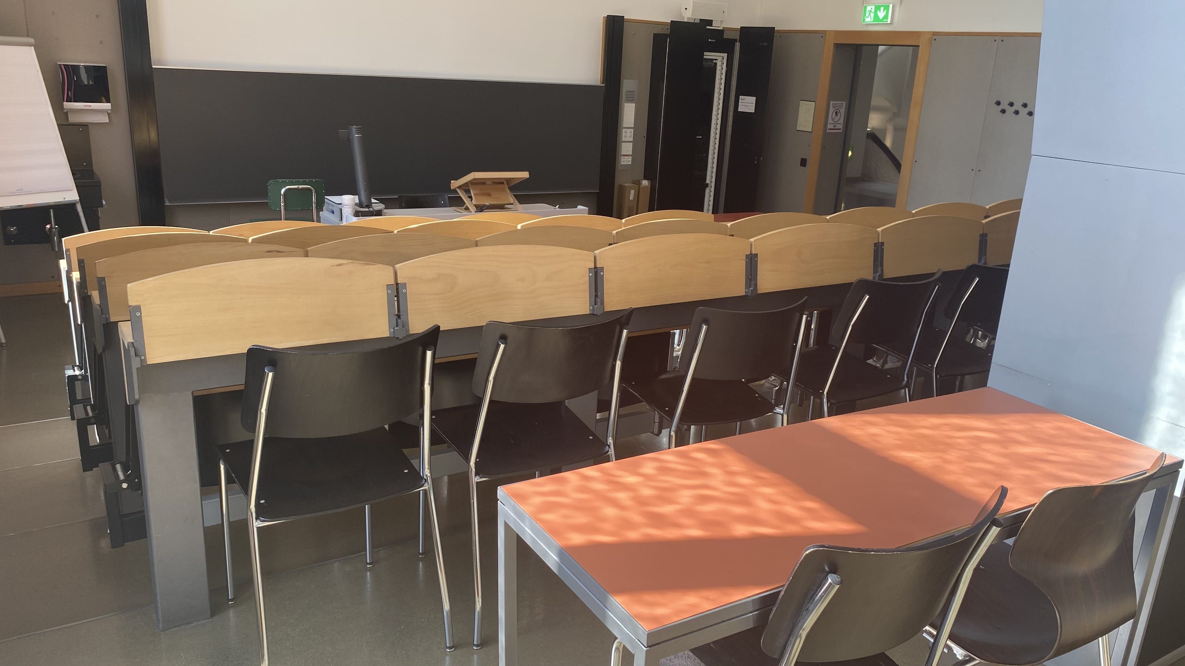 Hörsaal KOL-F-123: Ansicht zum Dozierendenpult hin. Die Eingangstür befindet sich rechts im Bild. In der hintersten Reihe befinden sich zwei Tische ohne fixe Bestuhlung.