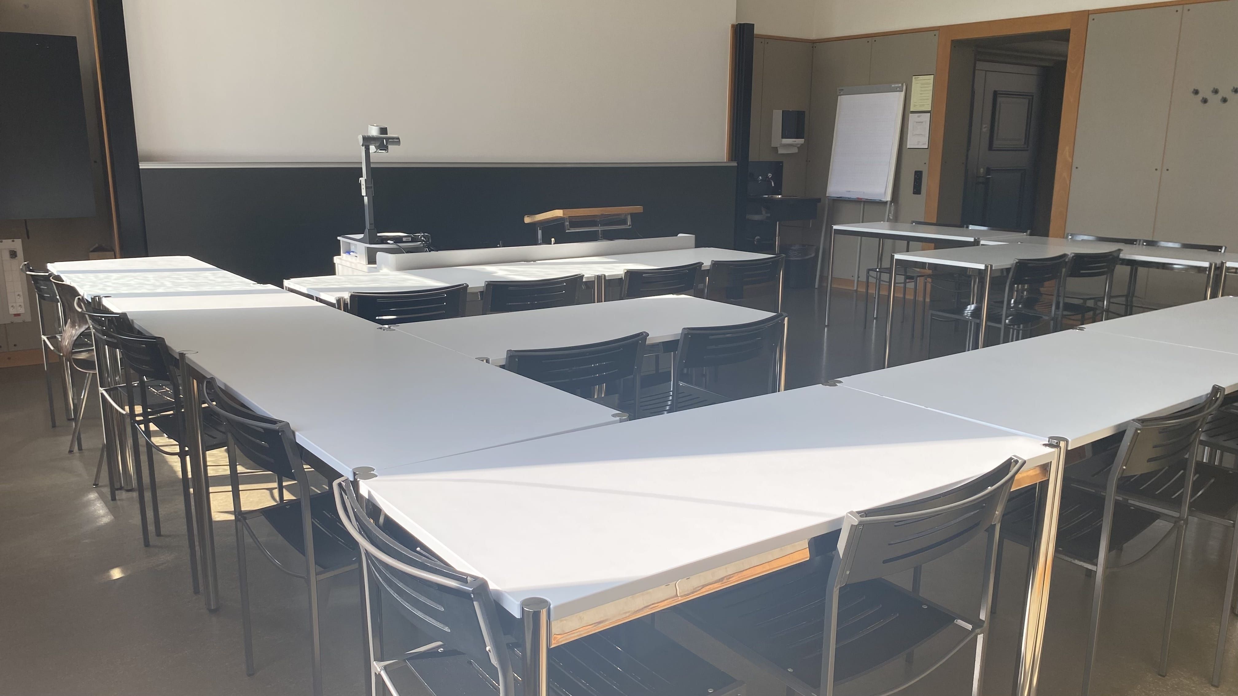 Seminarraum KOL-F-103: Ansicht auf den Raum zum Dozierendenpult hin. Die Eingangstür befindet sich rechts im Raum. Rechts von der Eingangstür befindet sich ein Lavabo.