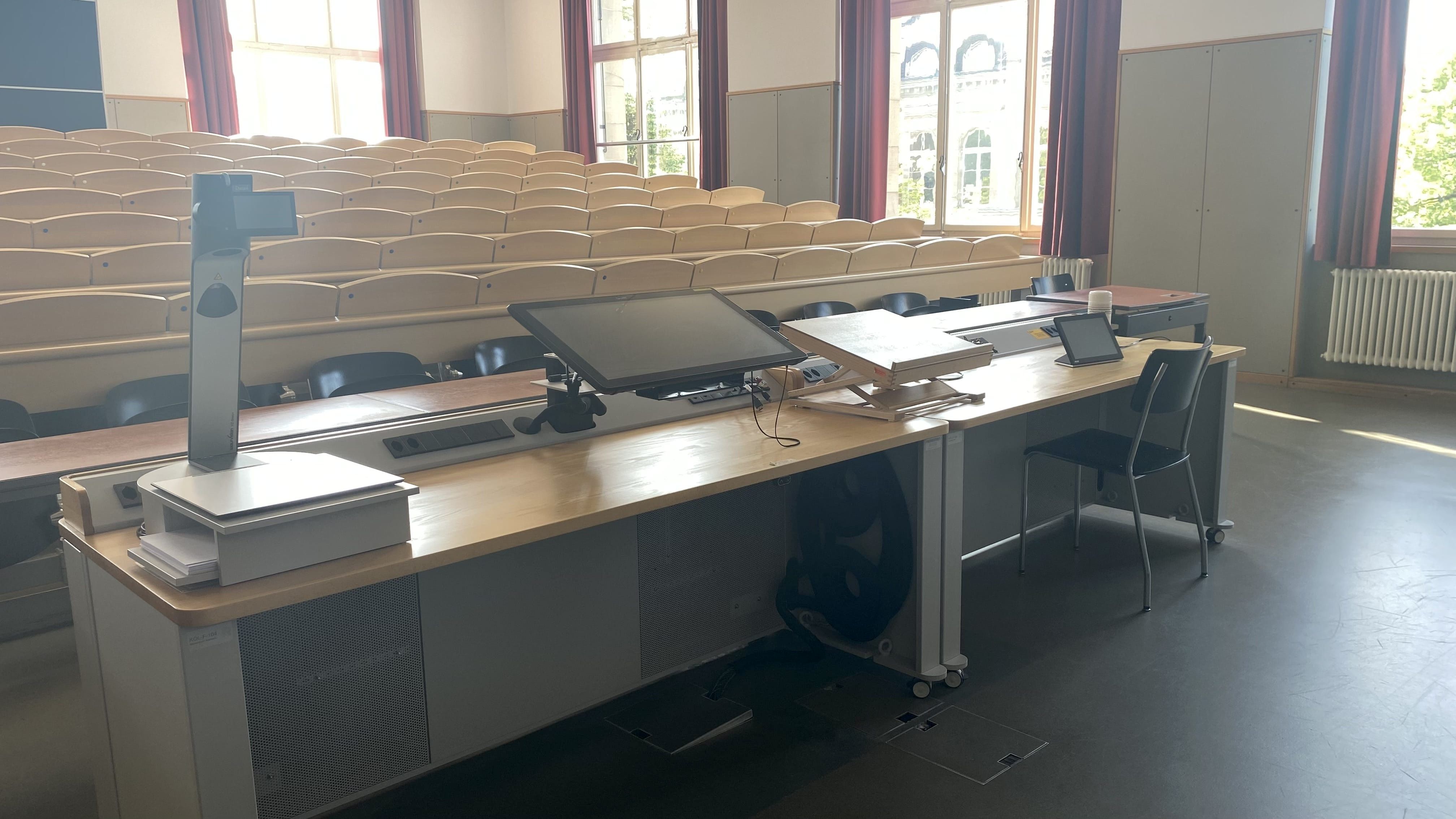 Hörsaal KOL-F-104: Ansicht auf das Dozierendenpult. Rechts vom Dozierendenpult befindet sich ein elektrisch höhenverstellbarer Tisch mit Rollstuhl-Signet.