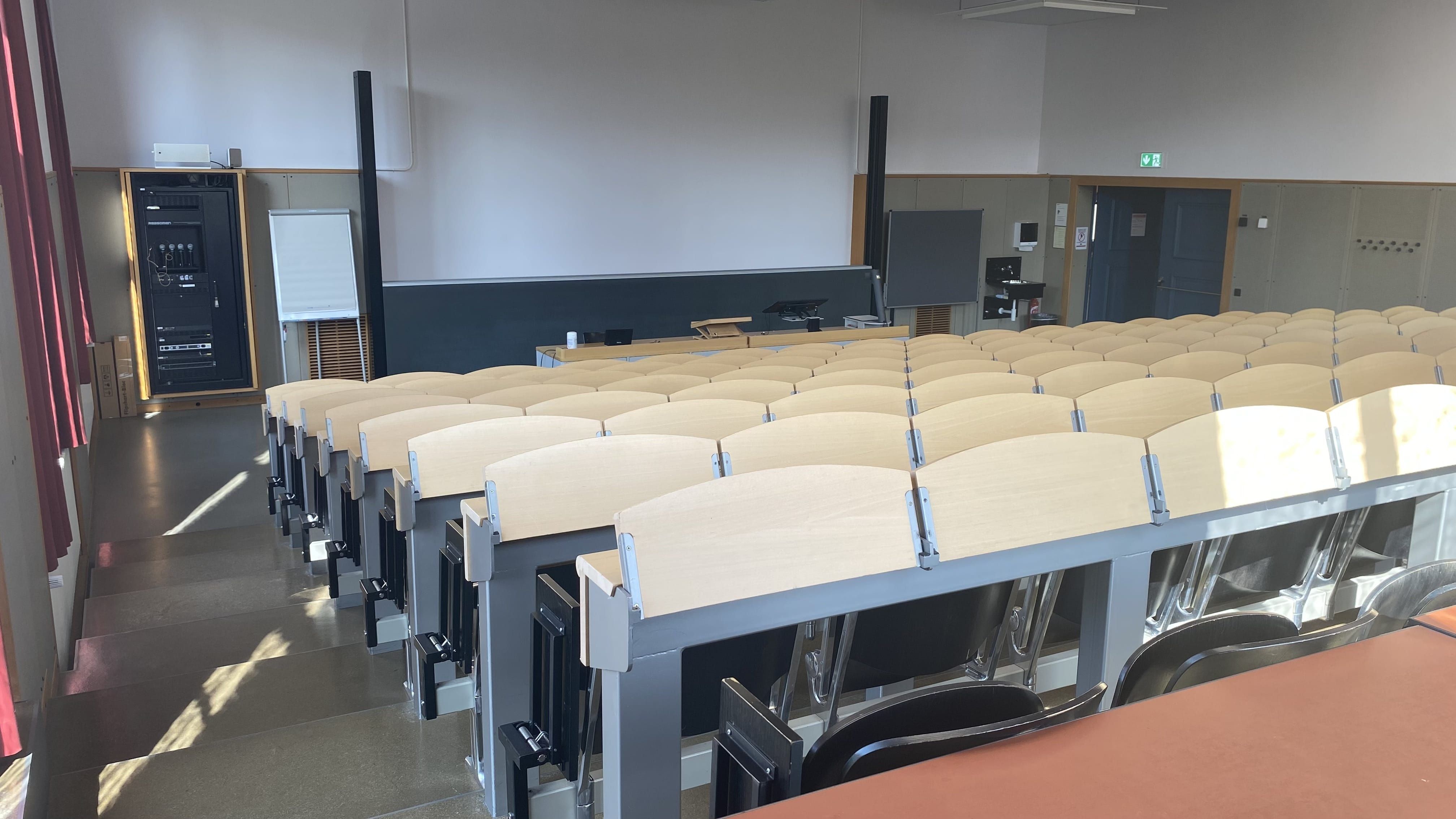 Hörsaal KOL-F-104: Ansicht zum Dozierndenpult hin. Die Eingangstür befindet sich rechts im Bild. Rechts neben der Eingangstür befindet sich ein Lavabo. Die Stufen links sind nicht mit Orientierungshilfen markiert.