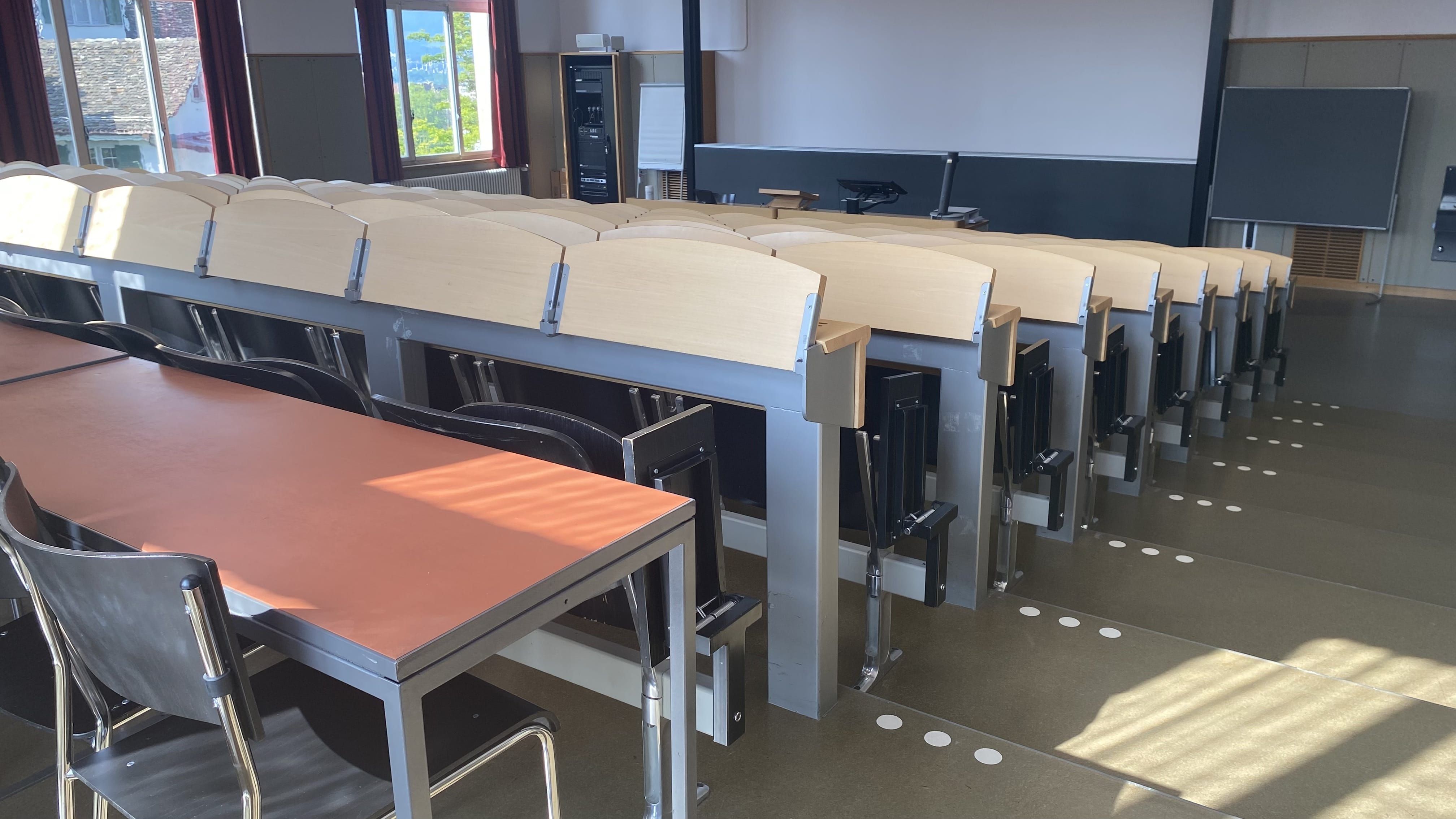 Hörsaal KOL-F-104: Ansicht zum Dozierendenpult hin. Die Stufen auf der rechten Seite des Raumes sind mit Orientierungshilfen markiert. Die hinterste Sitzreihe hat keine fixe Bestuhlung.