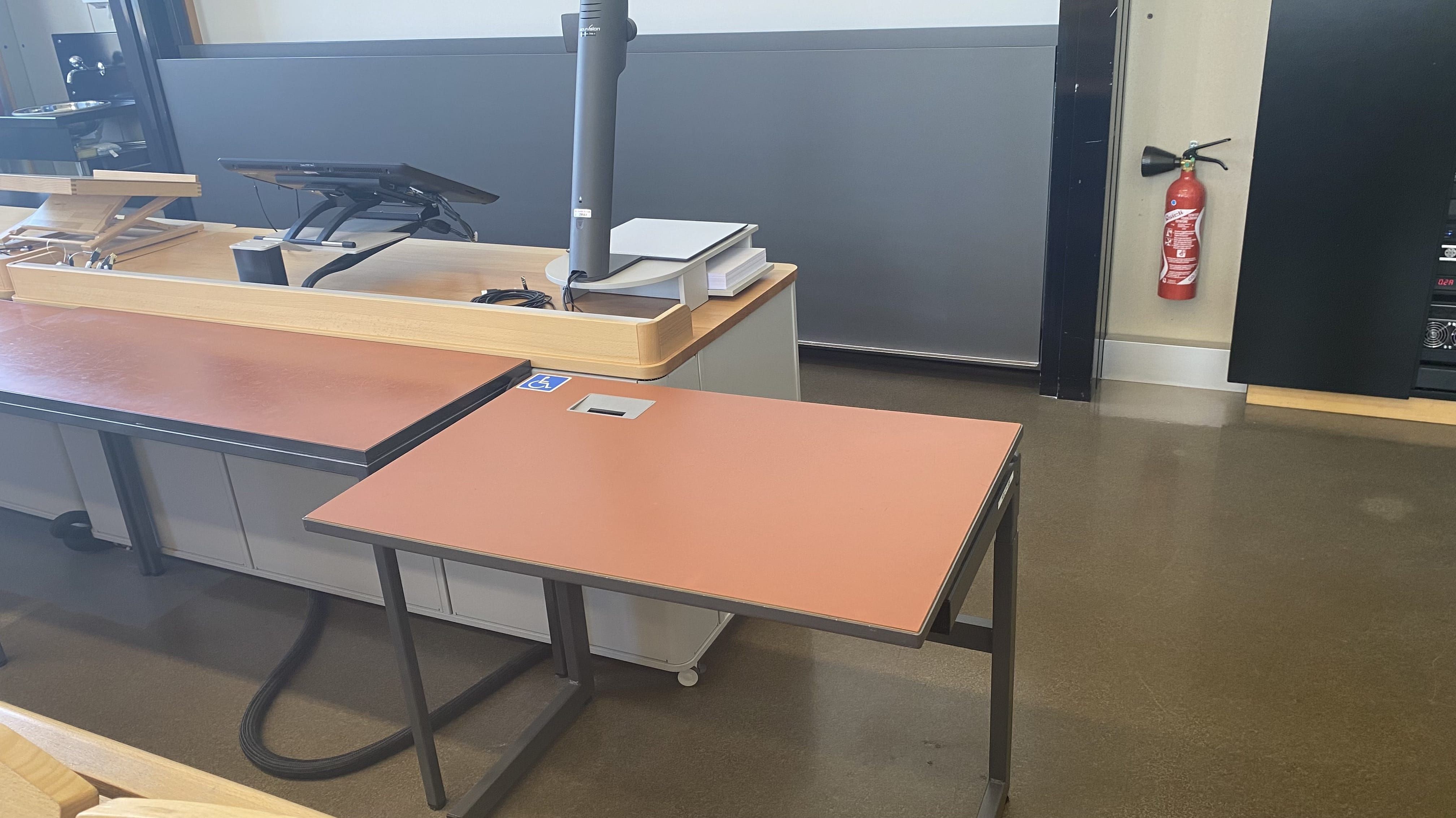 Hörsaal KOL-F-109: Ansicht auf den elektrisch höhenverstellbaren Tisch mit Rollstuhl-Signet. Der Tisch befindet sich in der vordersten Reihe auf der rechten Seite nahe der Eingangstür.