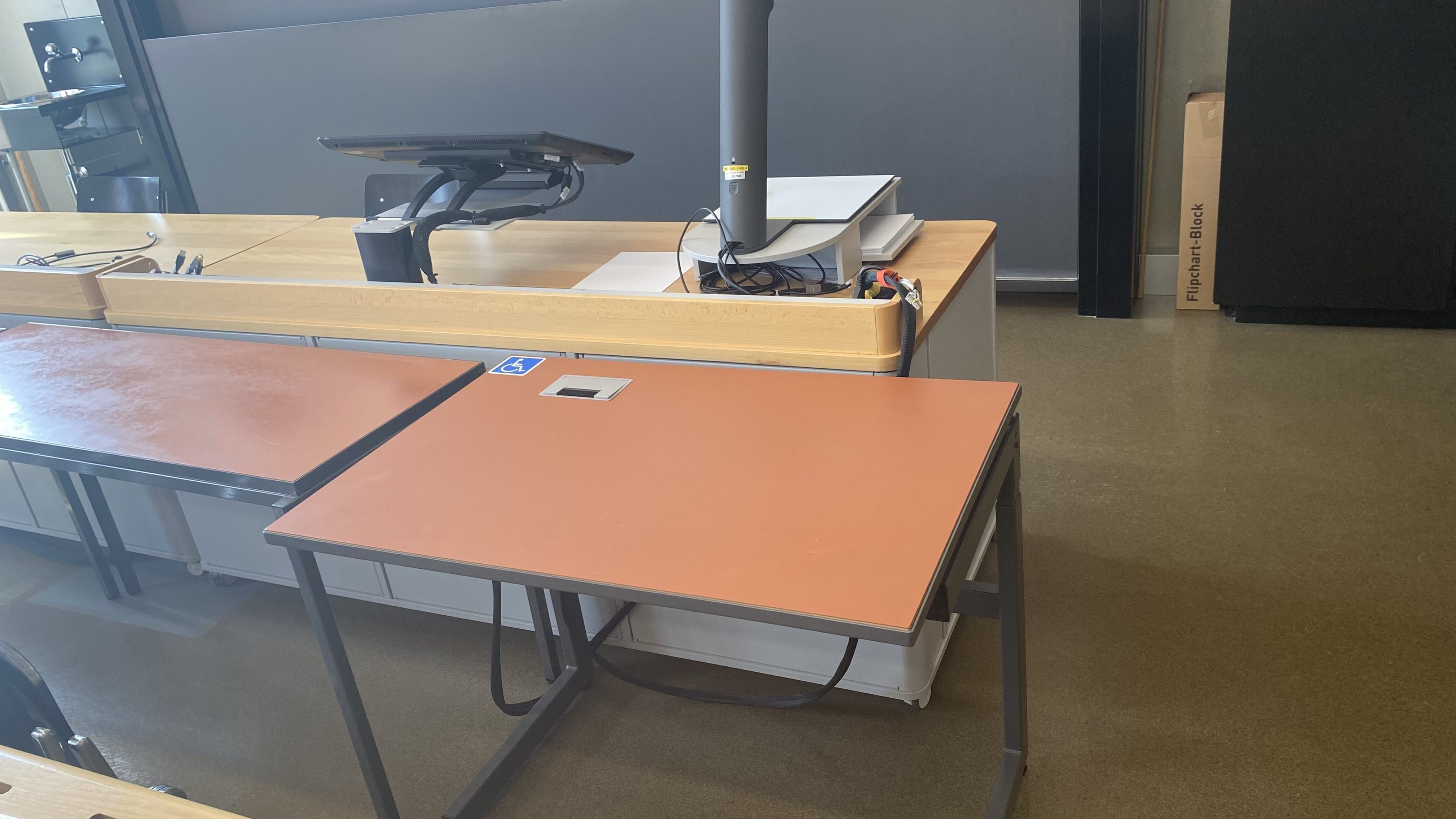 Hörsaal KOL-G-221: Ansicht auf den elektrisch höhenverstellbaren Tisch mit Rollstuhl-Signet. Der Tisch befindet sich in der vordersten Reihe rechts, nahe der Eingangstür.
