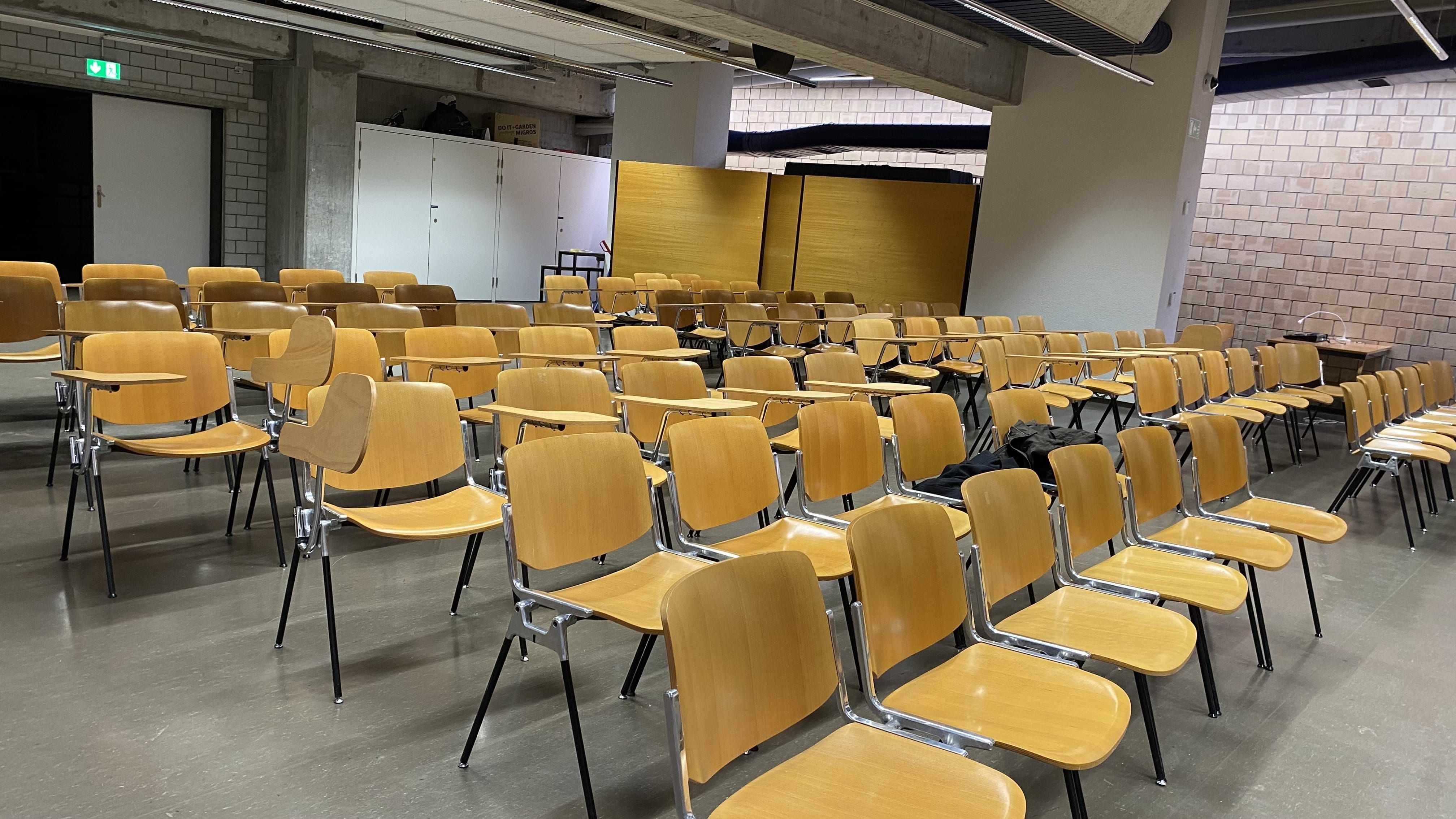 Aula Y21-F-65: Ansicht auf die Sitzreihen in der Aula. Es gibt Stühle mit und ohne fixe Tische.
