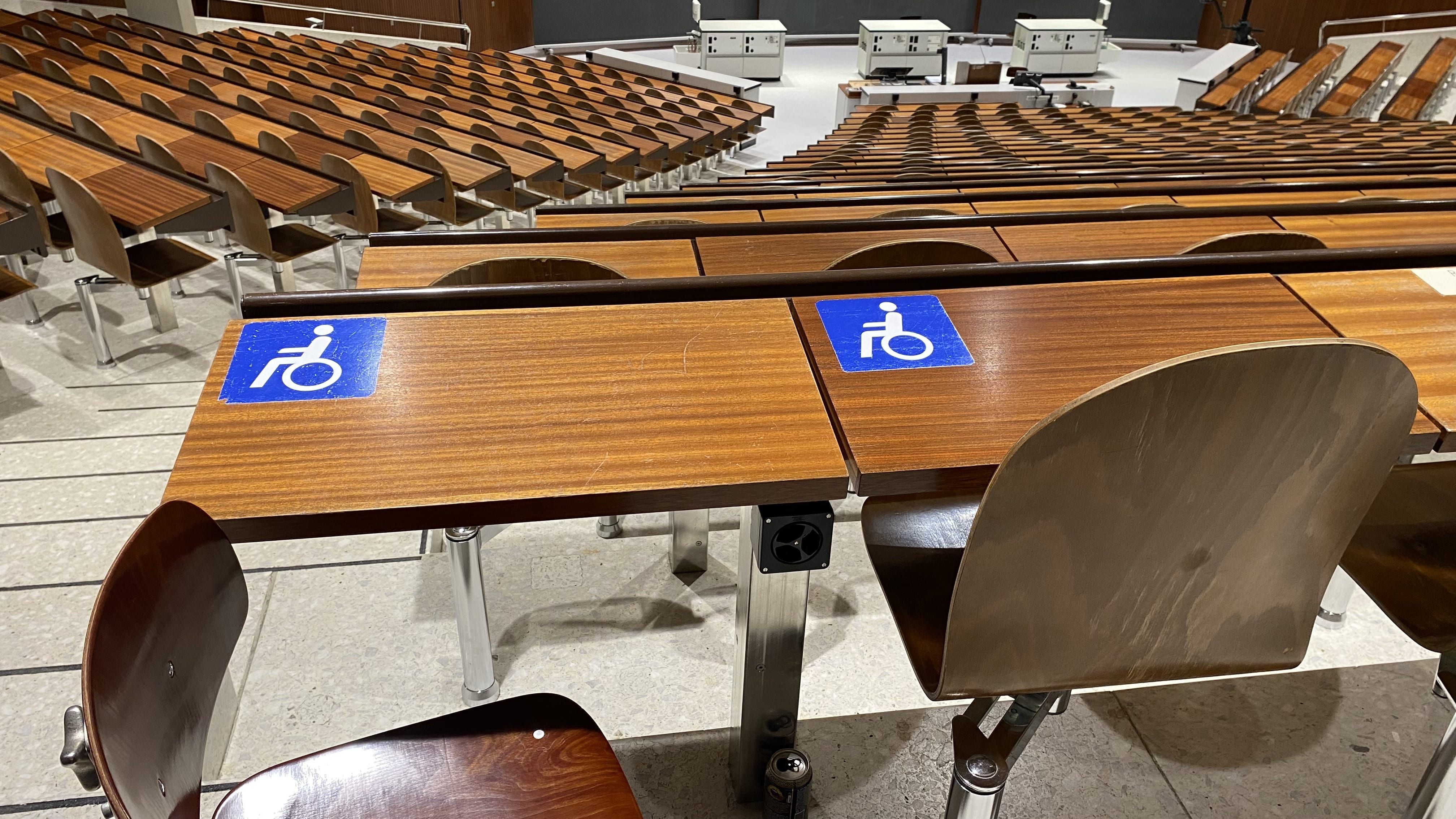 Hörsaal Y04-G-30: Ansicht auf die zwei von vier Tischen mit Rollstuhl-Signet ohne fixe Bestuhlung. Die Tische befinden sich in der hintersten, mittleren Sitzreihe links am Rand.