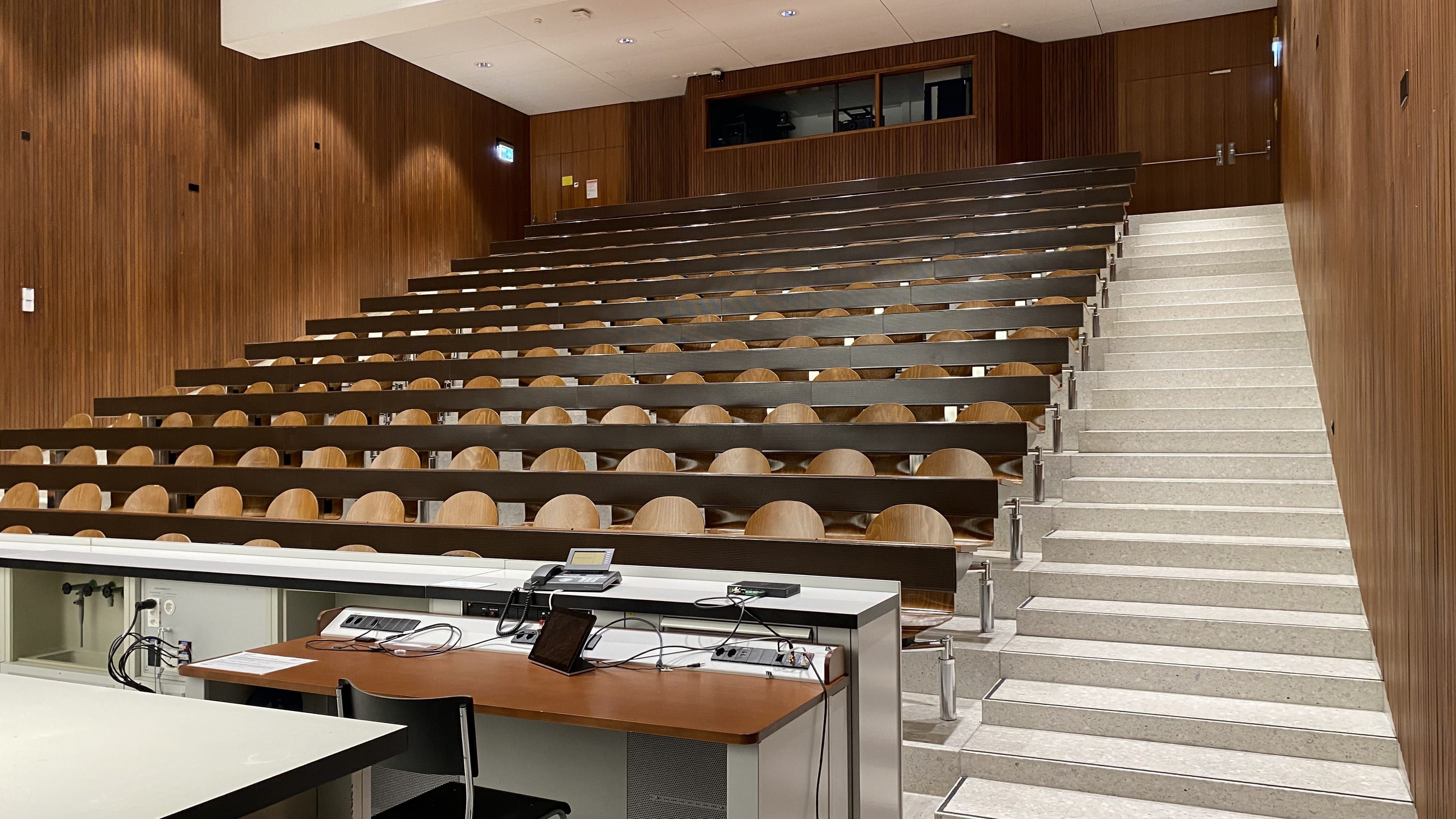 Hörsaal Y15-G-19: Ansicht auf dem Raum vom Dozierendenpult aus. Links und rechts oben sind Eingangstüren ersichtlich. Nur bei der rechten Türe befindet sich ein Tisch ohne fixe Bestuhlung.