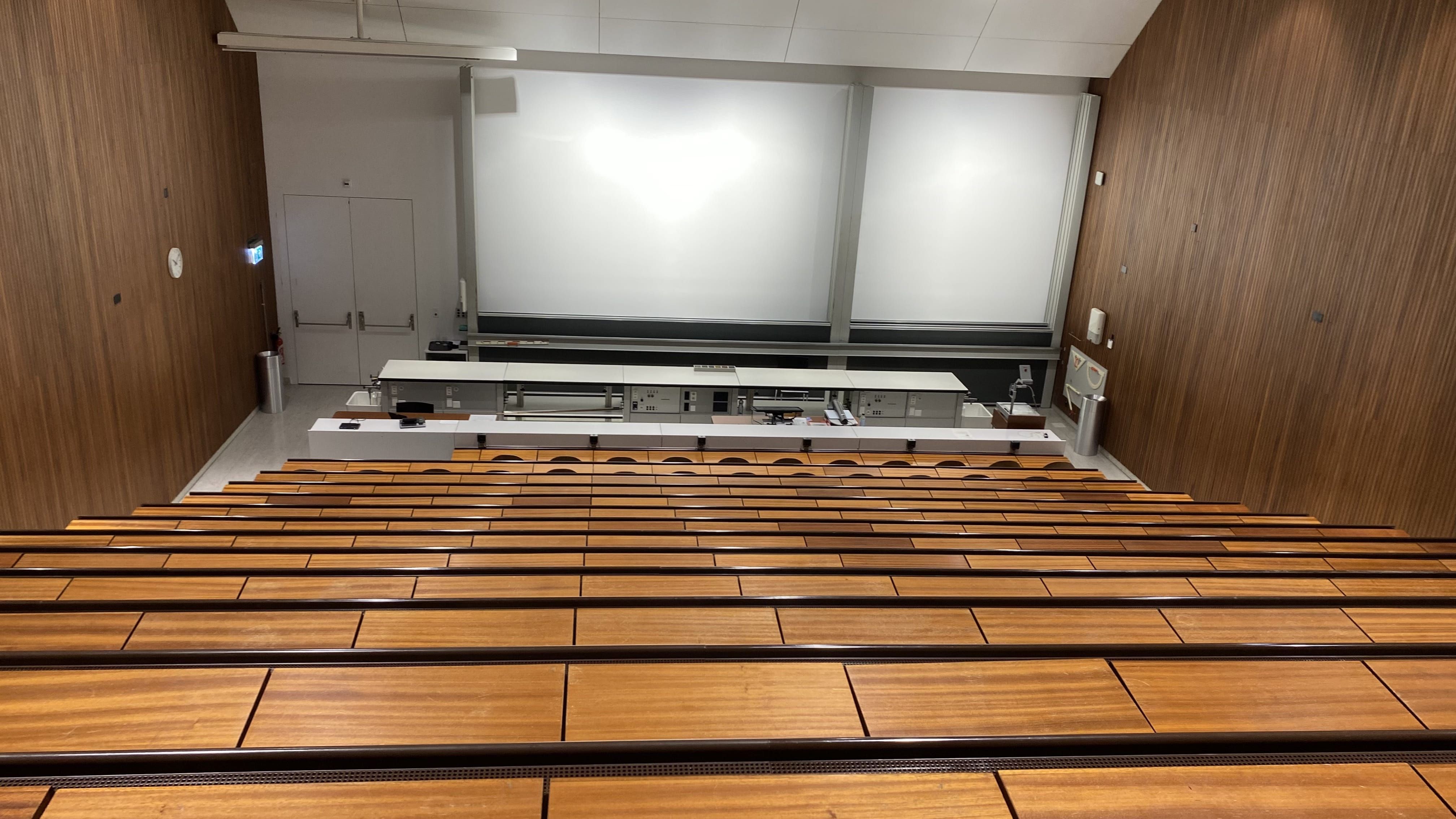 Hörsaal Y15-G-19: Ansicht aus der hintersten Reihe zum Dozierendenpult hin. Links unten im Raum befindet sich der Notausgang.